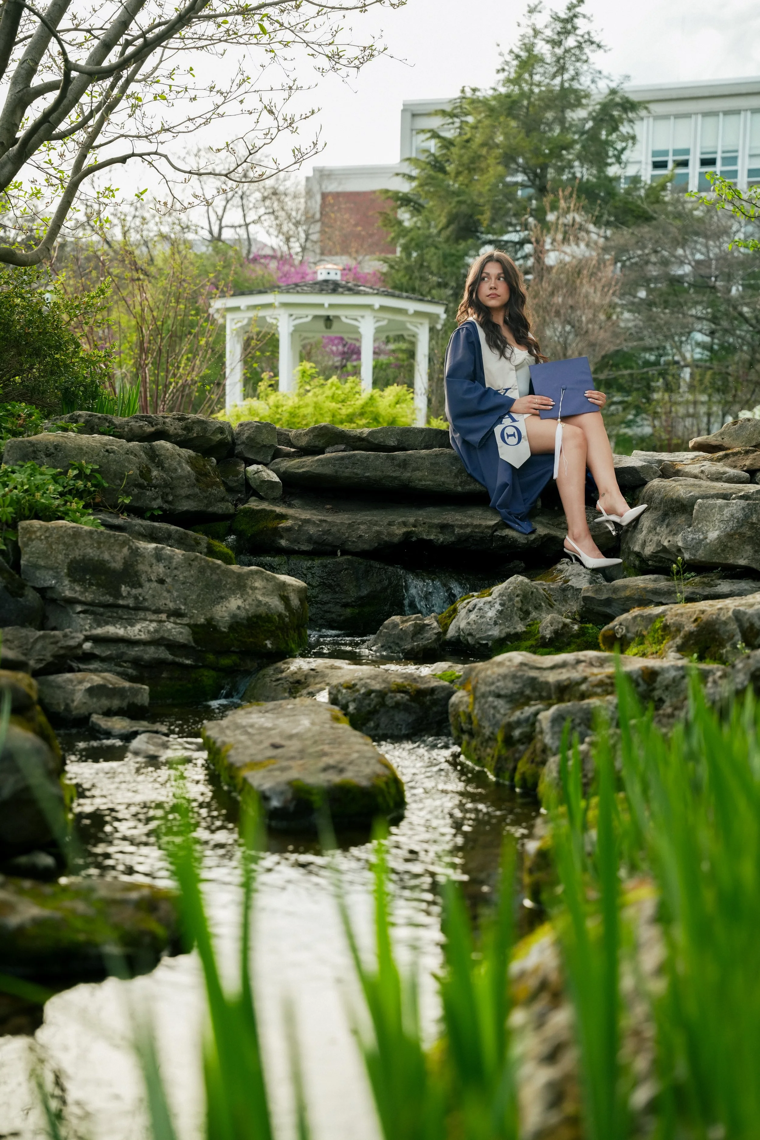 A young woman in a graduation gown sitting on rocks by a creek at a park.