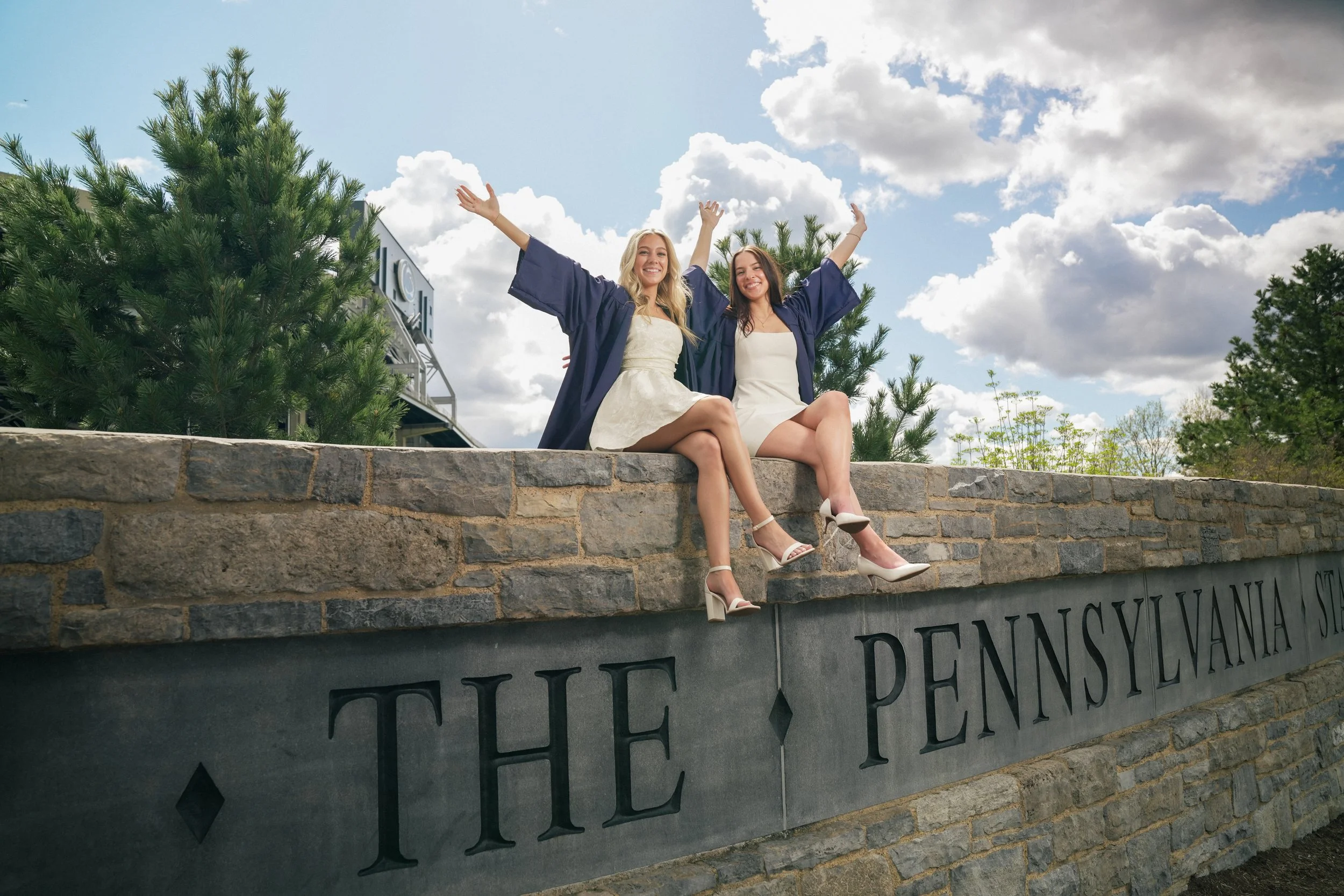 Two women in graduation caps and gowns sitting on a stone wall with a university sign, smiling with arms raised, under a partly cloudy sky.