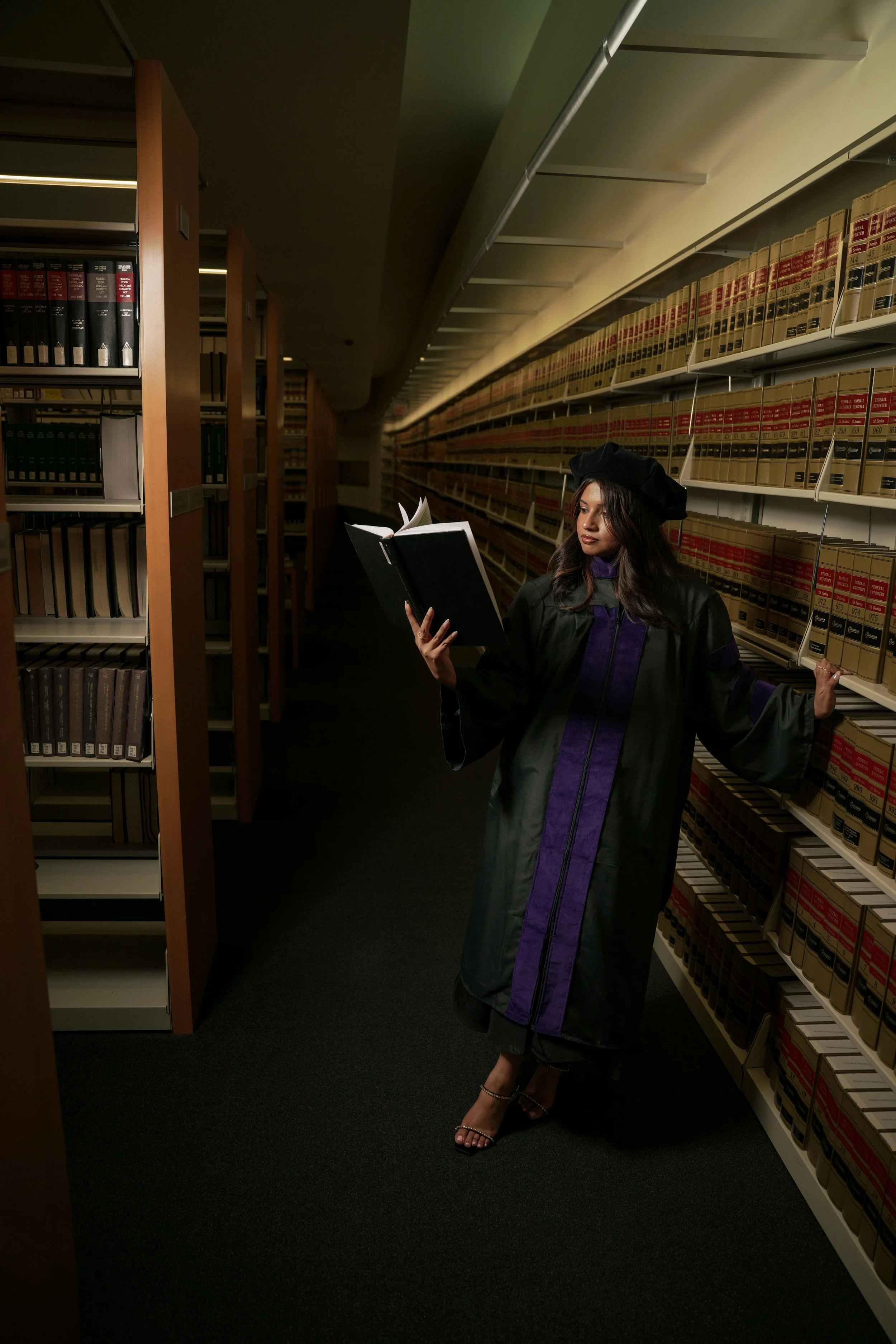 A woman dressed in a graduation gown and cap standing in a library aisle, reading a book, with shelves of books in the background.