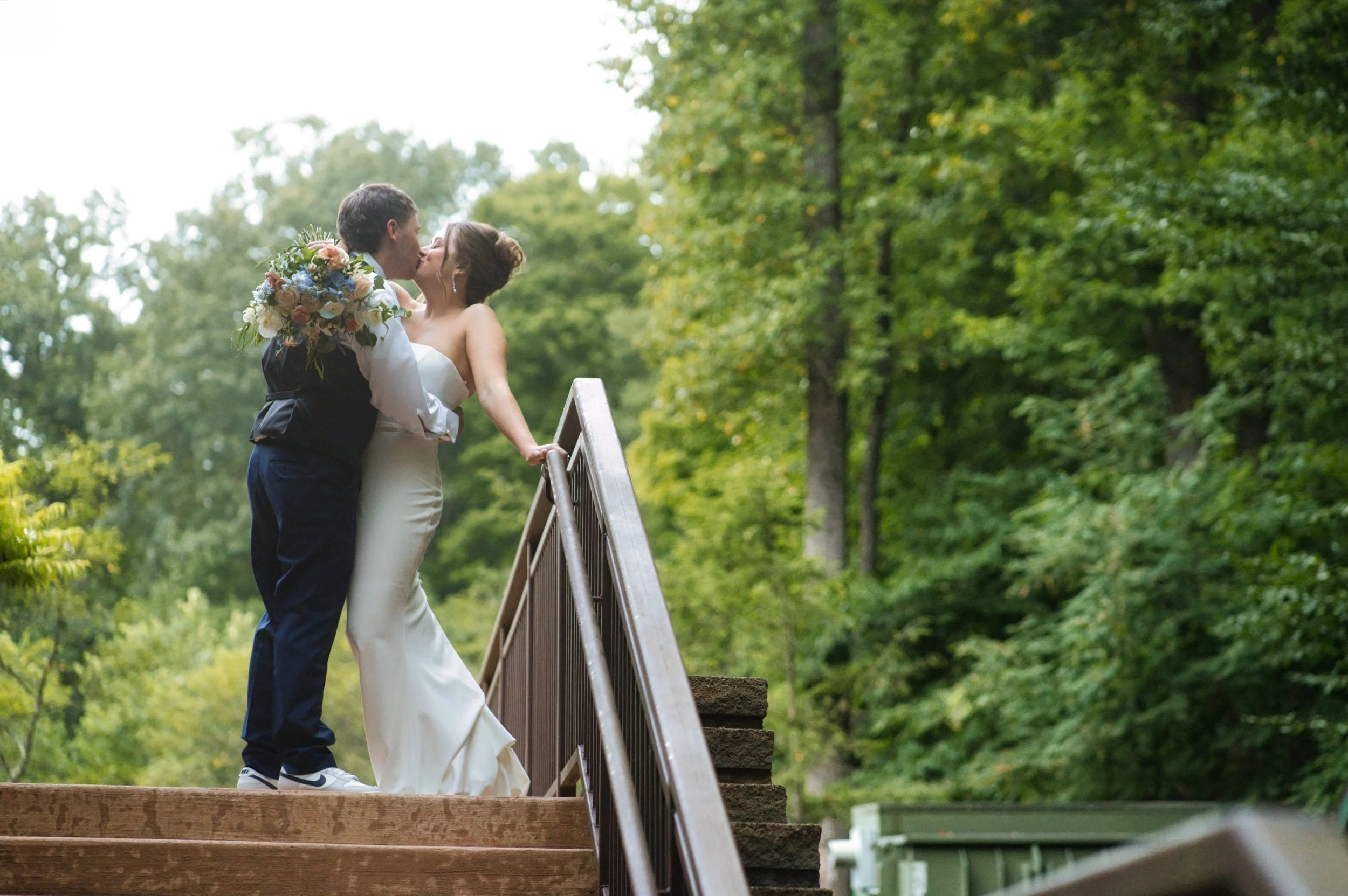 Bride and groom kissing on a wooden bridge in a green outdoor setting, with the bride holding a bouquet of flowers.