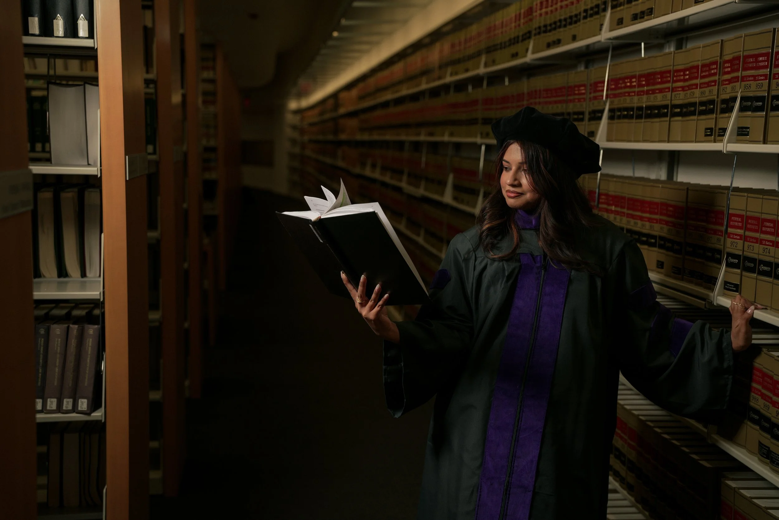 A young woman in a graduation gown and cap stands in the library aisle, reading a black folder with papers, surrounded by tall metal shelves filled with labeled file boxes.