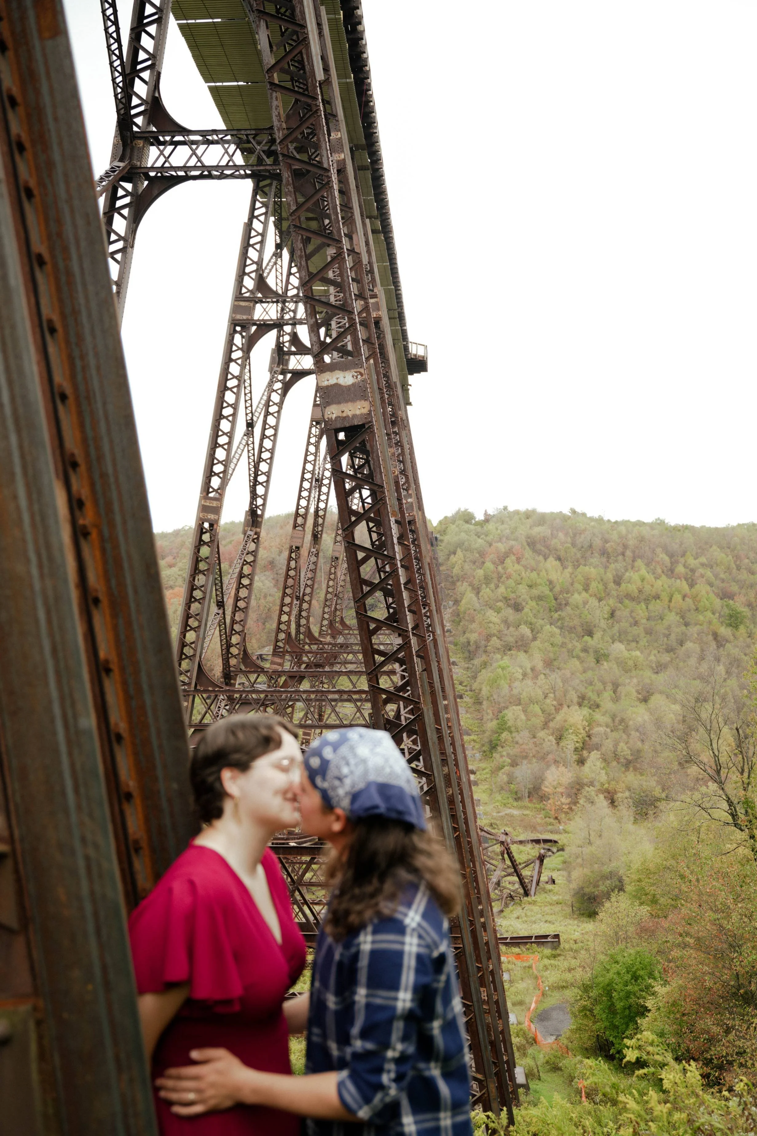 Two women sharing a kiss beneath a large, rusted, metal bridge structure in a rural, hilly area with trees and greenery in the background.
