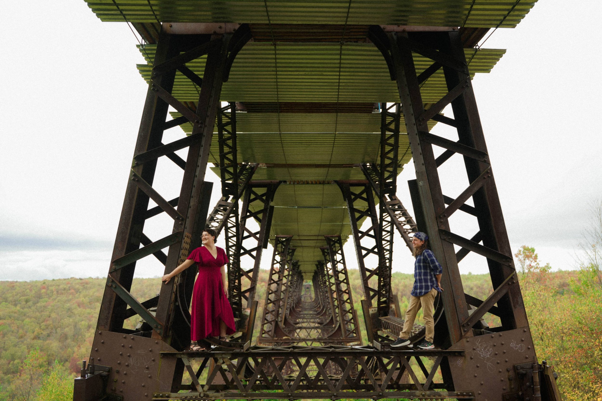 Two people standing underneath a large, rusted elevated bridge with a green metal deck, scenic fall foliage in the background.