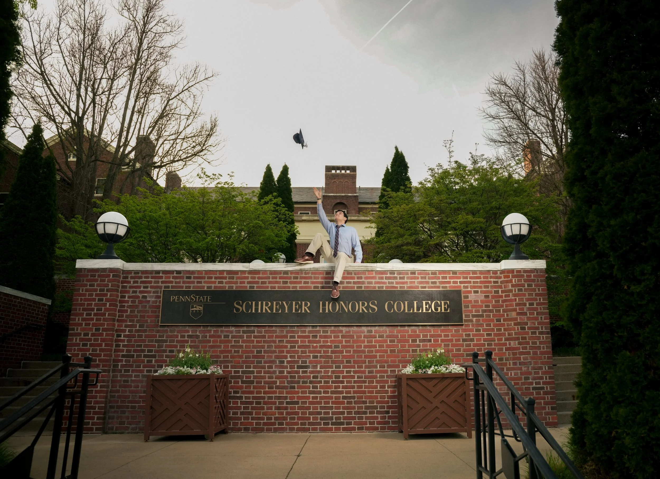 A young man in business attire sitting on a brick wall at Penn State Schreyer Honors College, tossing a graduation cap into the air outdoors on a cloudy day, with leafless and evergreen trees in the background.