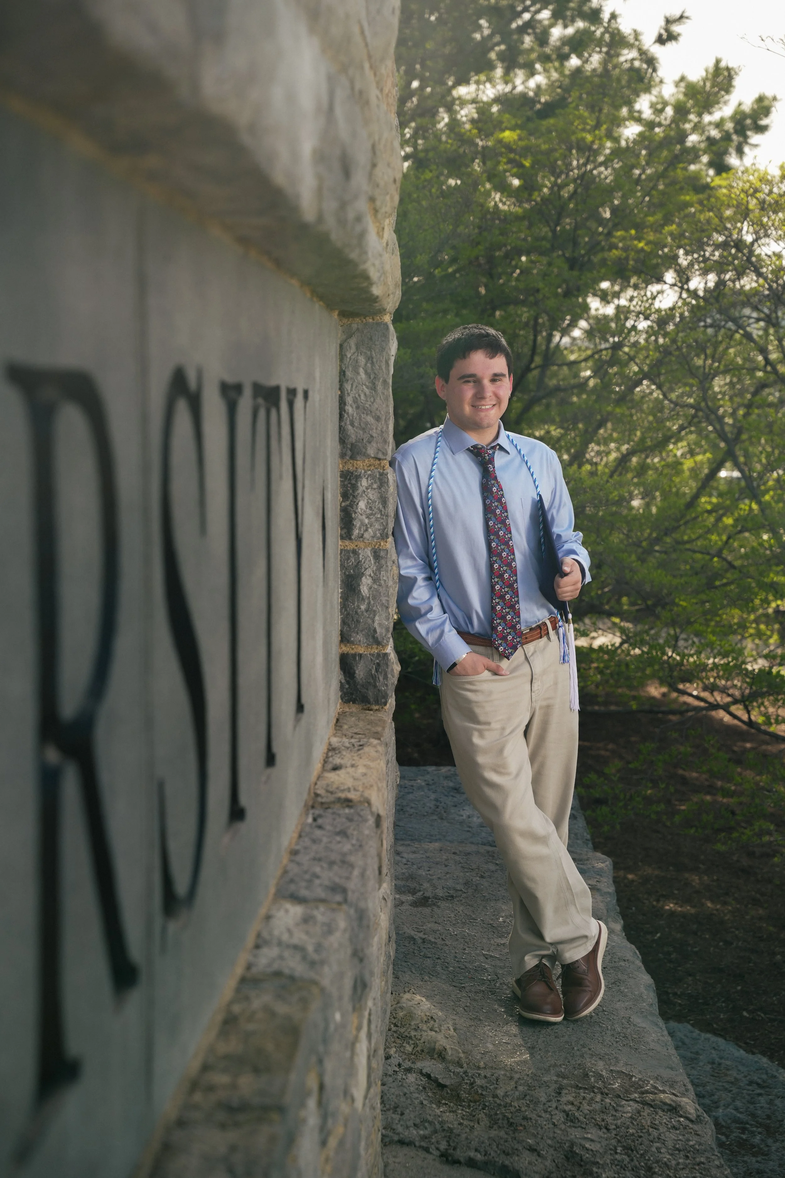 A young man in dress clothes leaning against a stone wall with trees in the background, smiling at the camera.