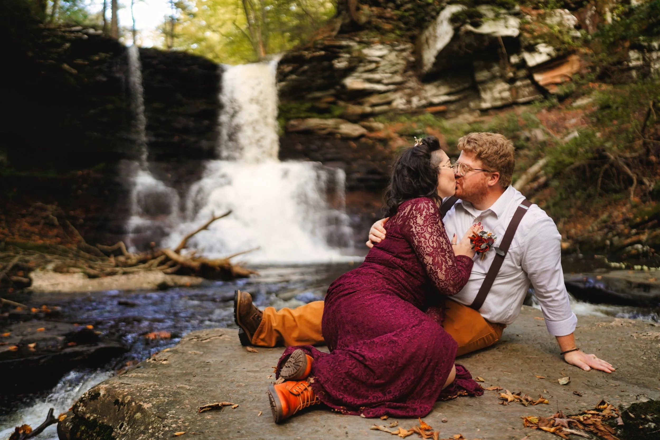A couple kissing on a rock by a waterfall in a forest setting, with autumn leaves scattered on the ground.