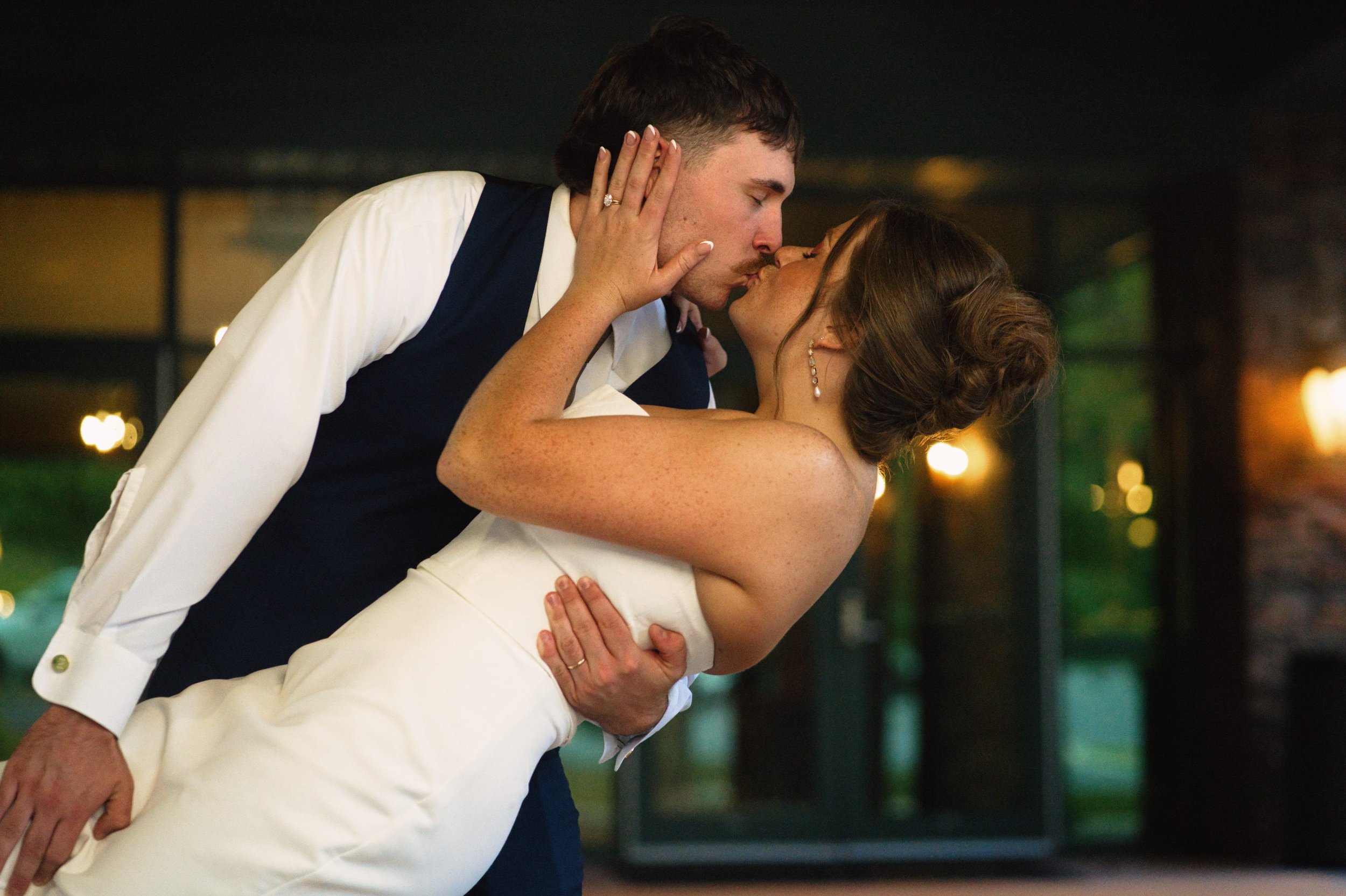 A newlywed couple kissing during their wedding reception, with the groom dipping the bride. The bride is dressed in a strapless white gown and those in a white dress shirt with a dark vest.