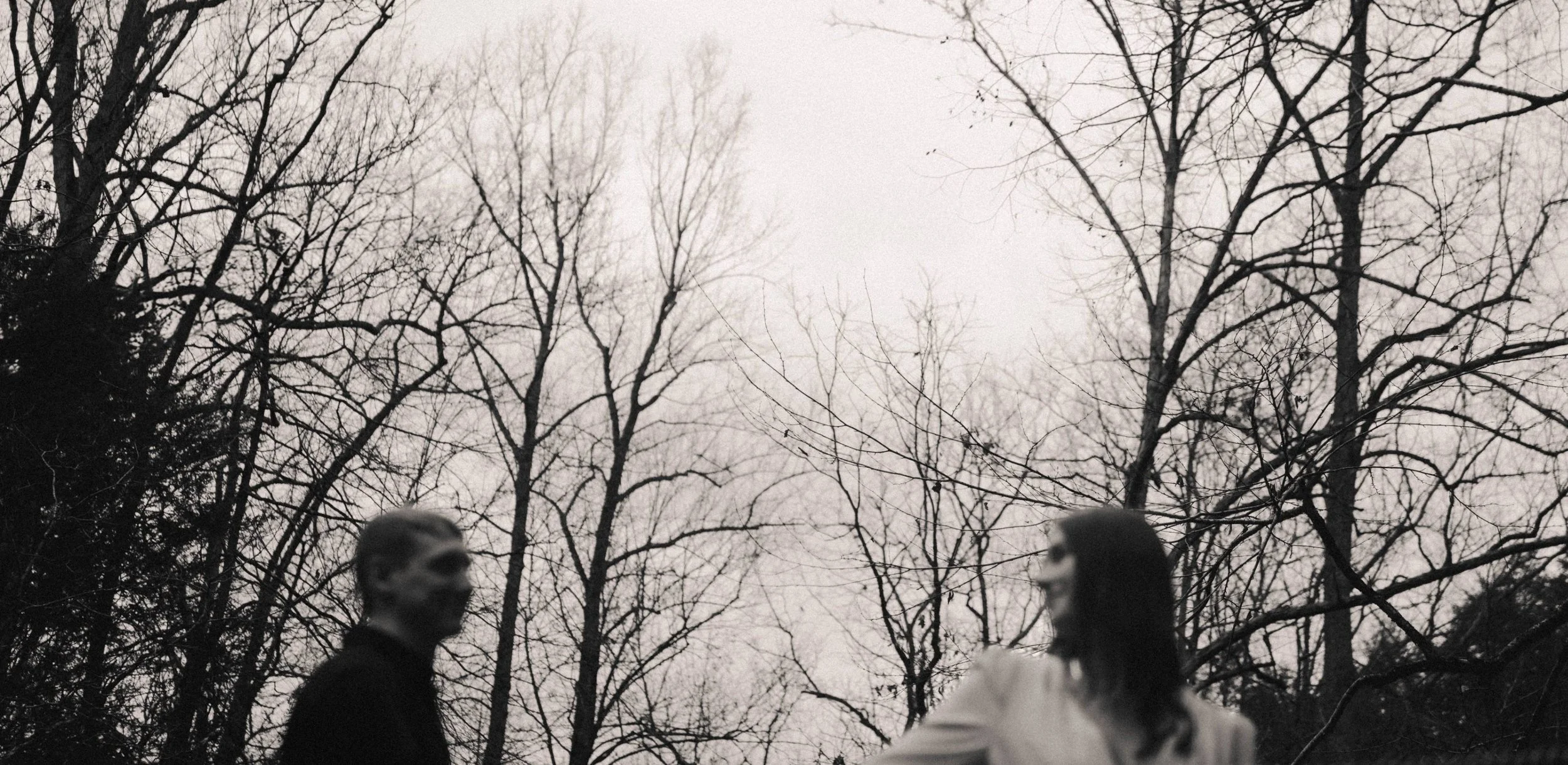 Two people wearing masks face each other outdoors among leafless trees on a cloudy day.