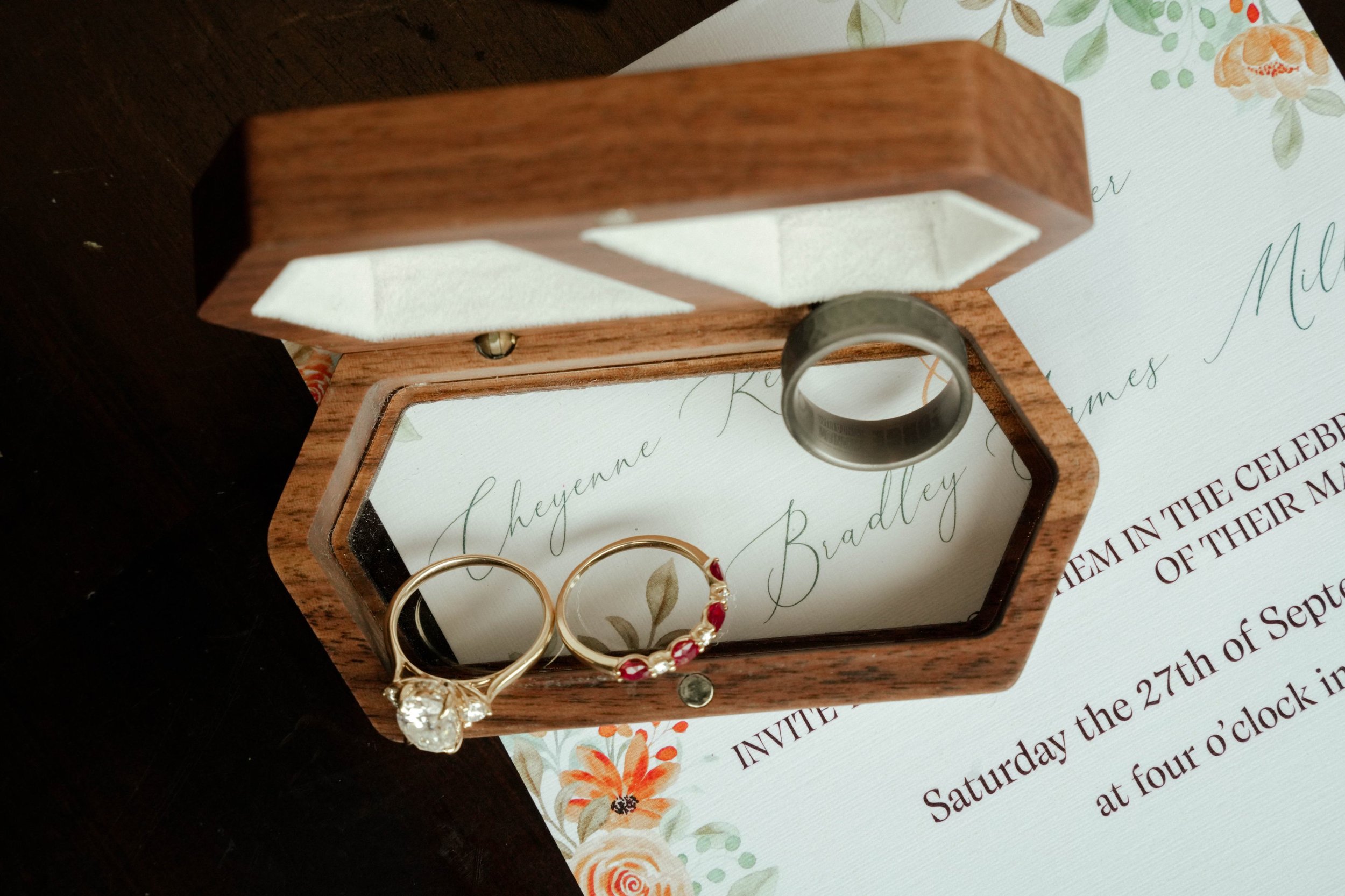 A wooden ring box with a mirror inside, holding two rings—one with a large clear stone and another with embedded red and white stones—placed above a wedding invitation with floral decorations and cursive writing.