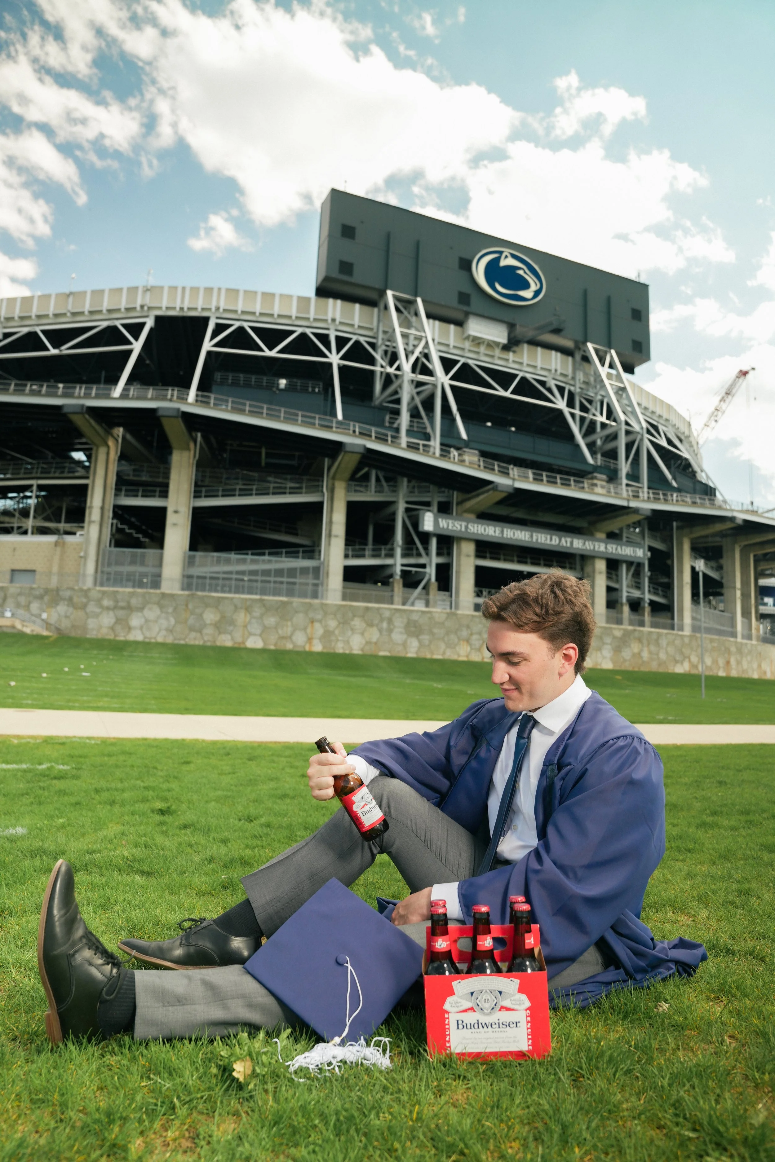 A young man in a suit and tie, wearing a blue jacket, gray pants, and black shoes, sitting on the grass outside a stadium with a logo and a sign indicating it is Beaver Stadium. He is holding a beer bottle, with several more in a red case next to him