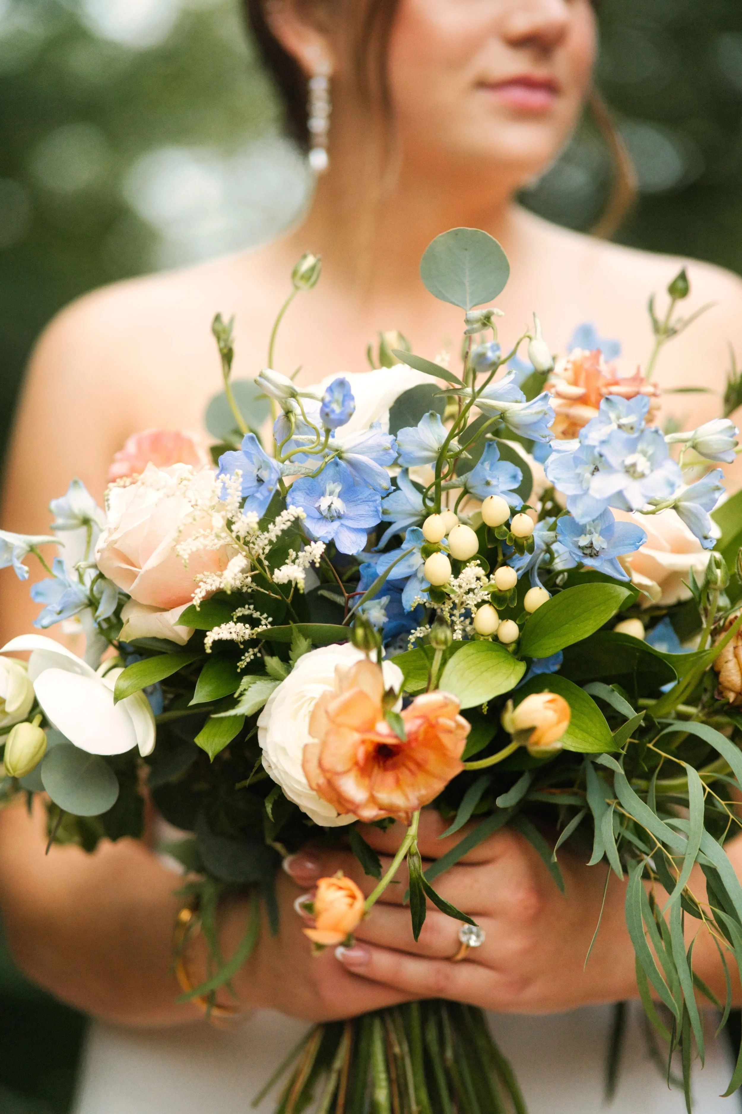 A woman holding a colorful bouquet of flowers, including roses, delphiniums, and berries, outdoors with a blurred green background.