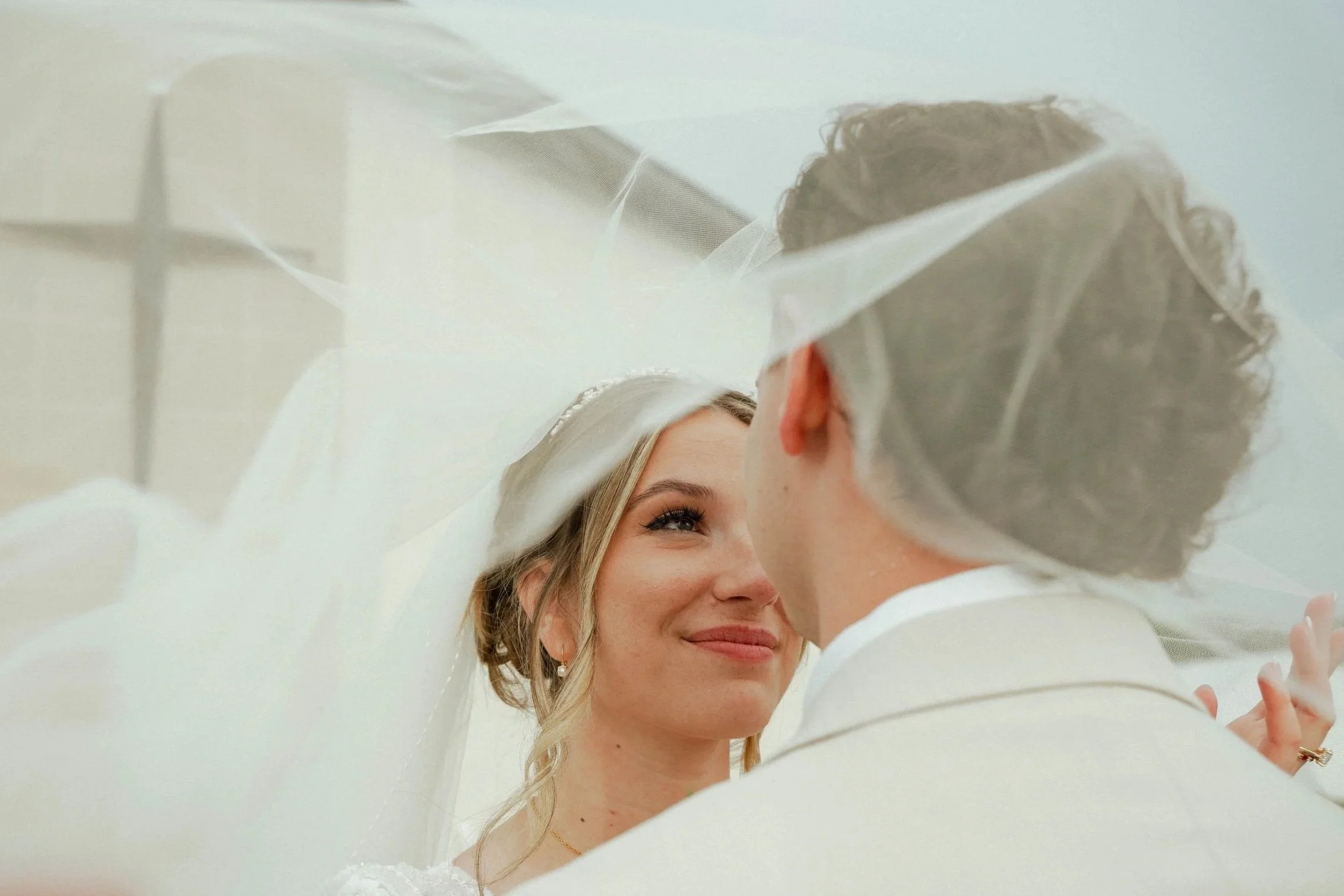 A bride and groom share a close moment beneath a wedding veil, looking at each other with affection.