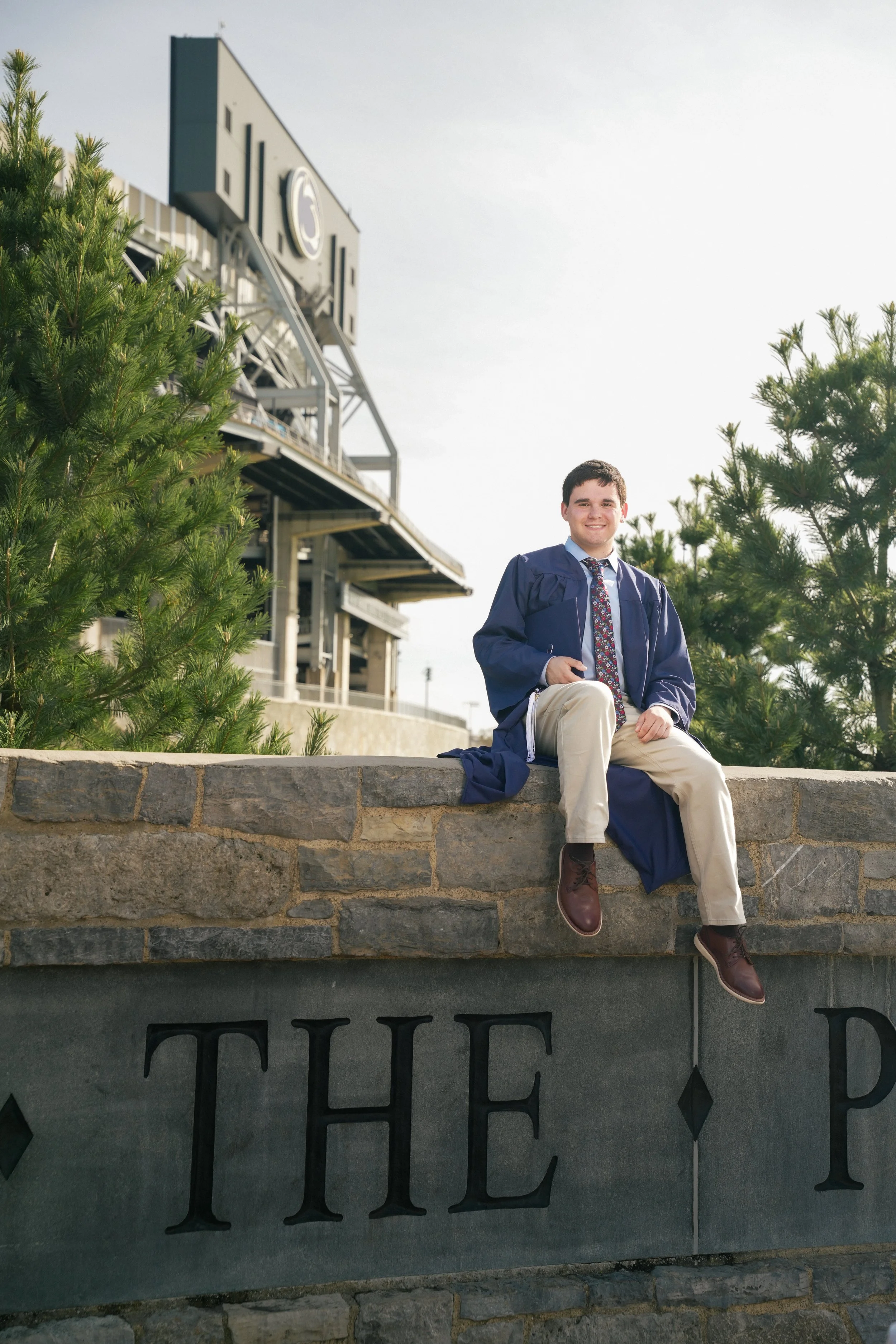 Young man in graduation cap and gown sitting on a stone ledge with trees behind him and a building in the background.