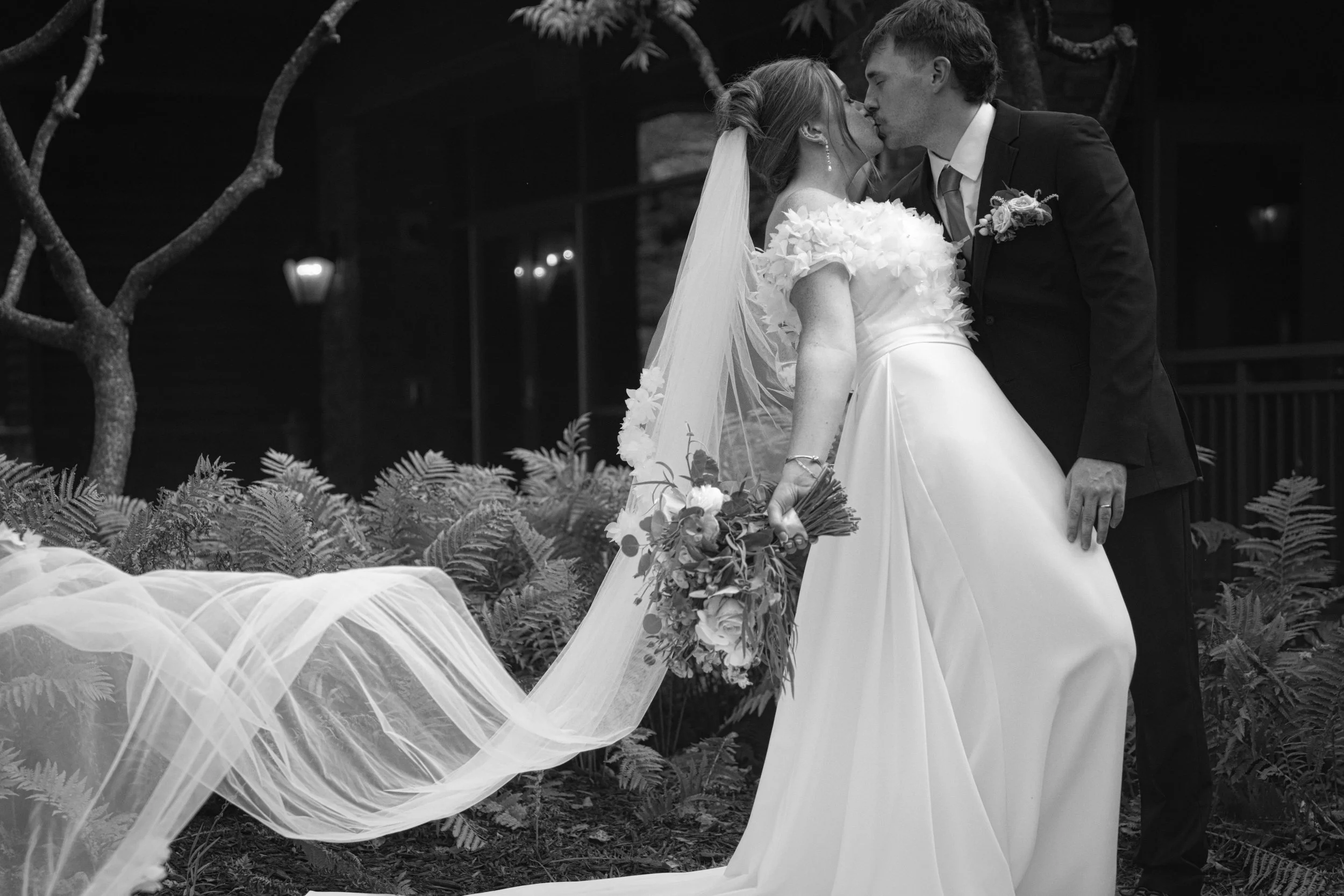 A black and white photo of a bride and groom kissing outdoors, with the bride wearing a white wedding gown and veil, holding a bouquet, and the groom wearing a black suit and tie, surrounded by ferns and trees.