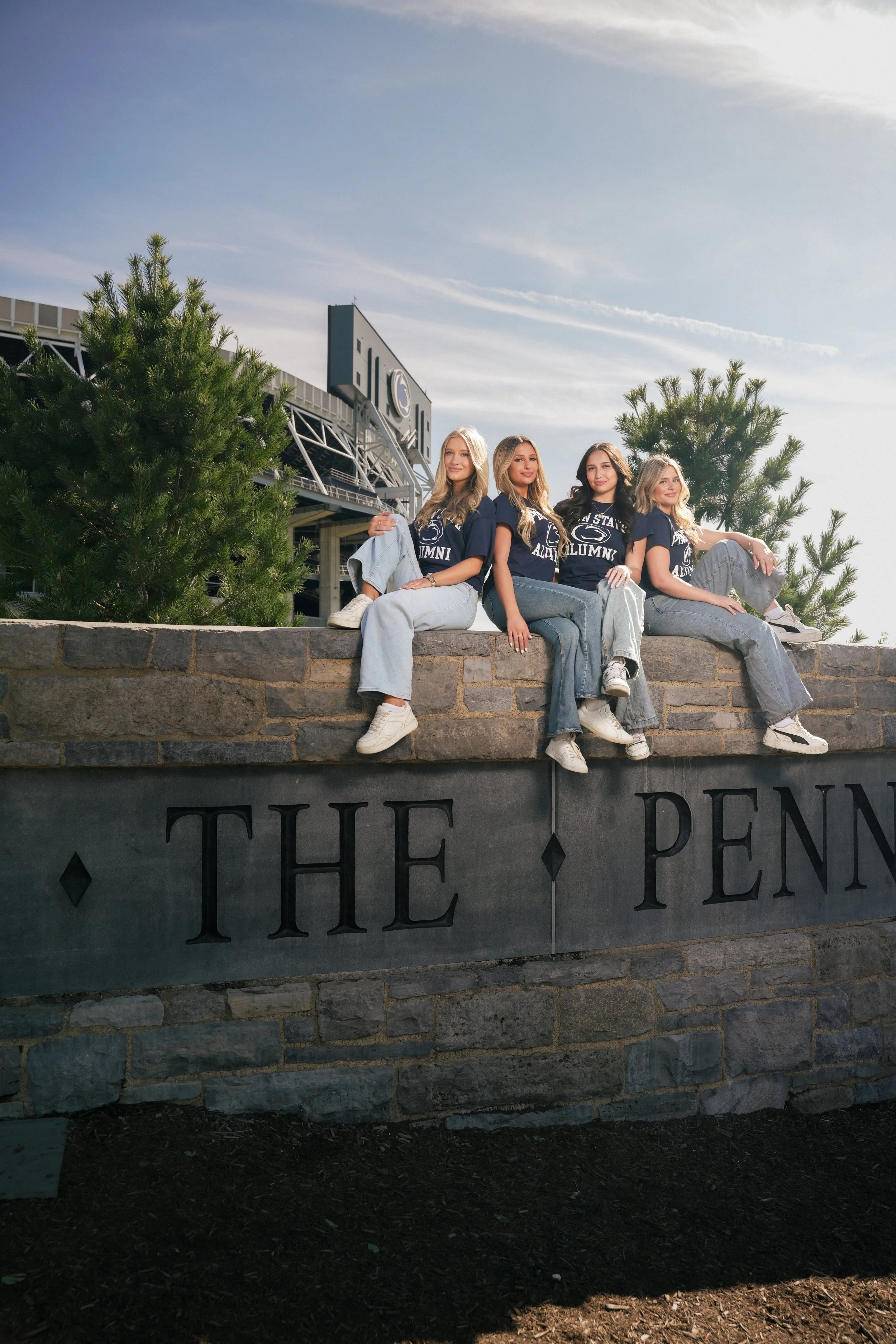 Four women sitting on a stone wall in front of a sign that reads 'The Penn State University Campus' with trees and a building in the background.