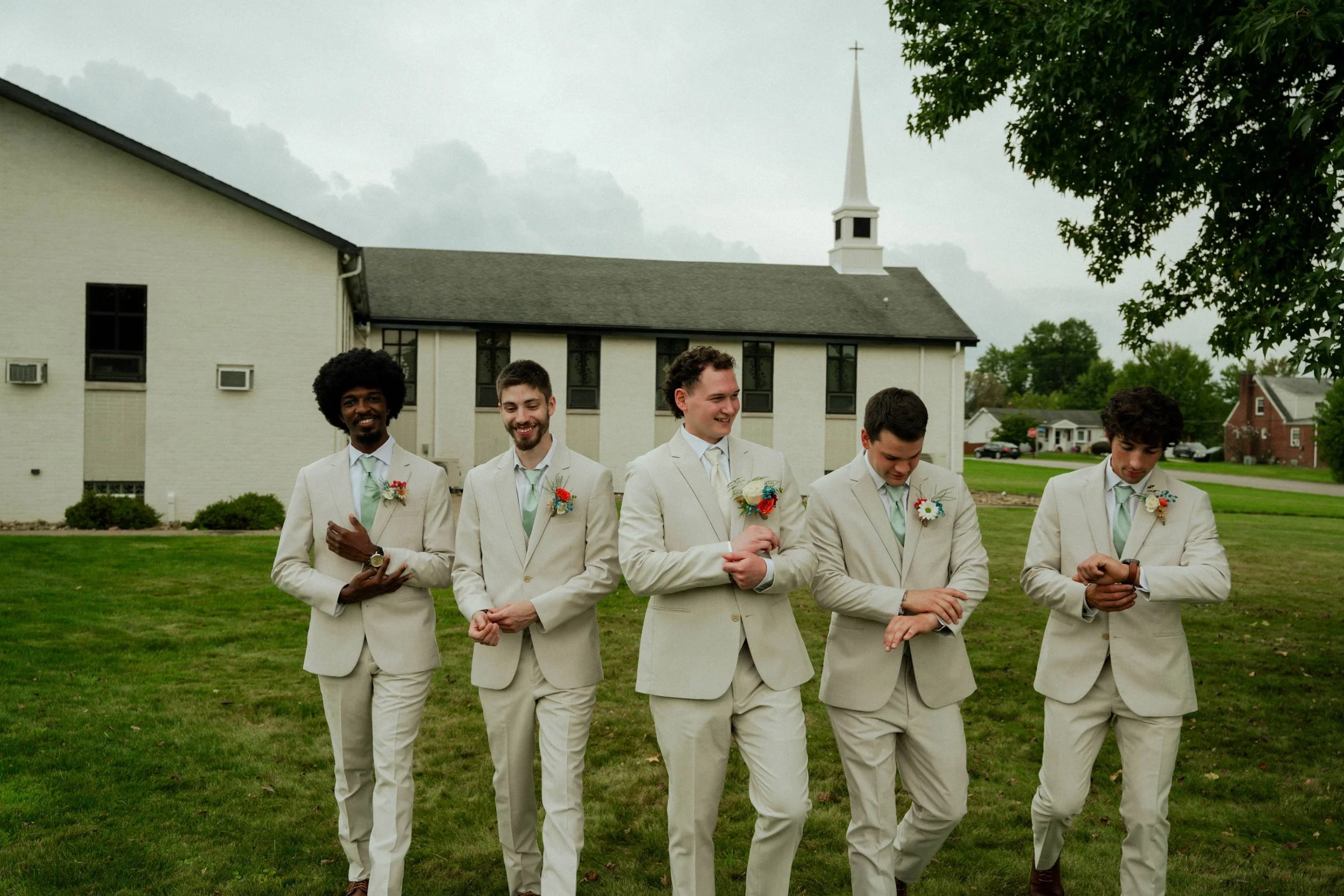 Group of five men in beige suits walking outdoors in front of a church with a steeple, during a wedding celebration, smiling and looking relaxed.