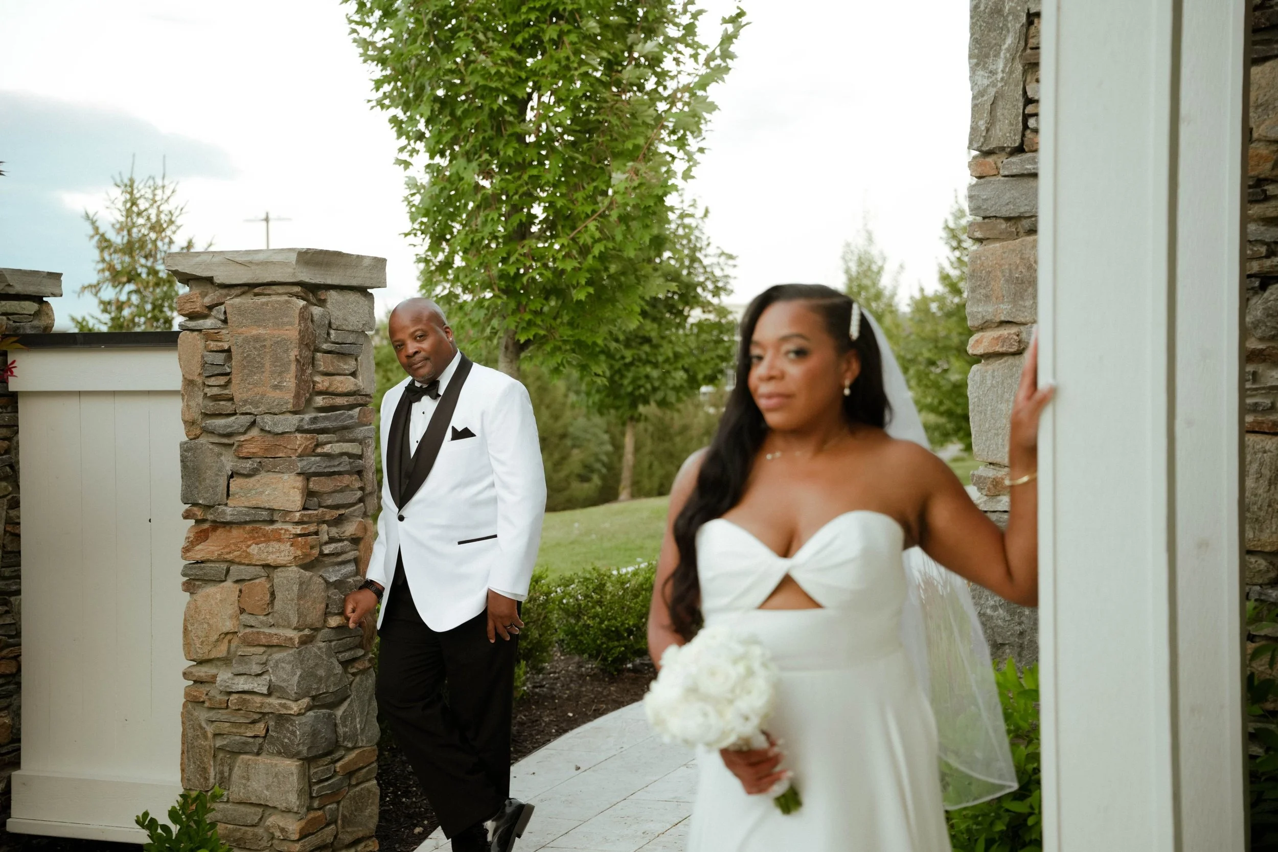 Bride in a strapless white wedding gown holding a bouquet of white flowers stands outdoors near a stone wall. Groom in a white tuxedo jacket with black lapels, black pants, and bow tie stands nearby, looking at her, in a garden setting with green tre