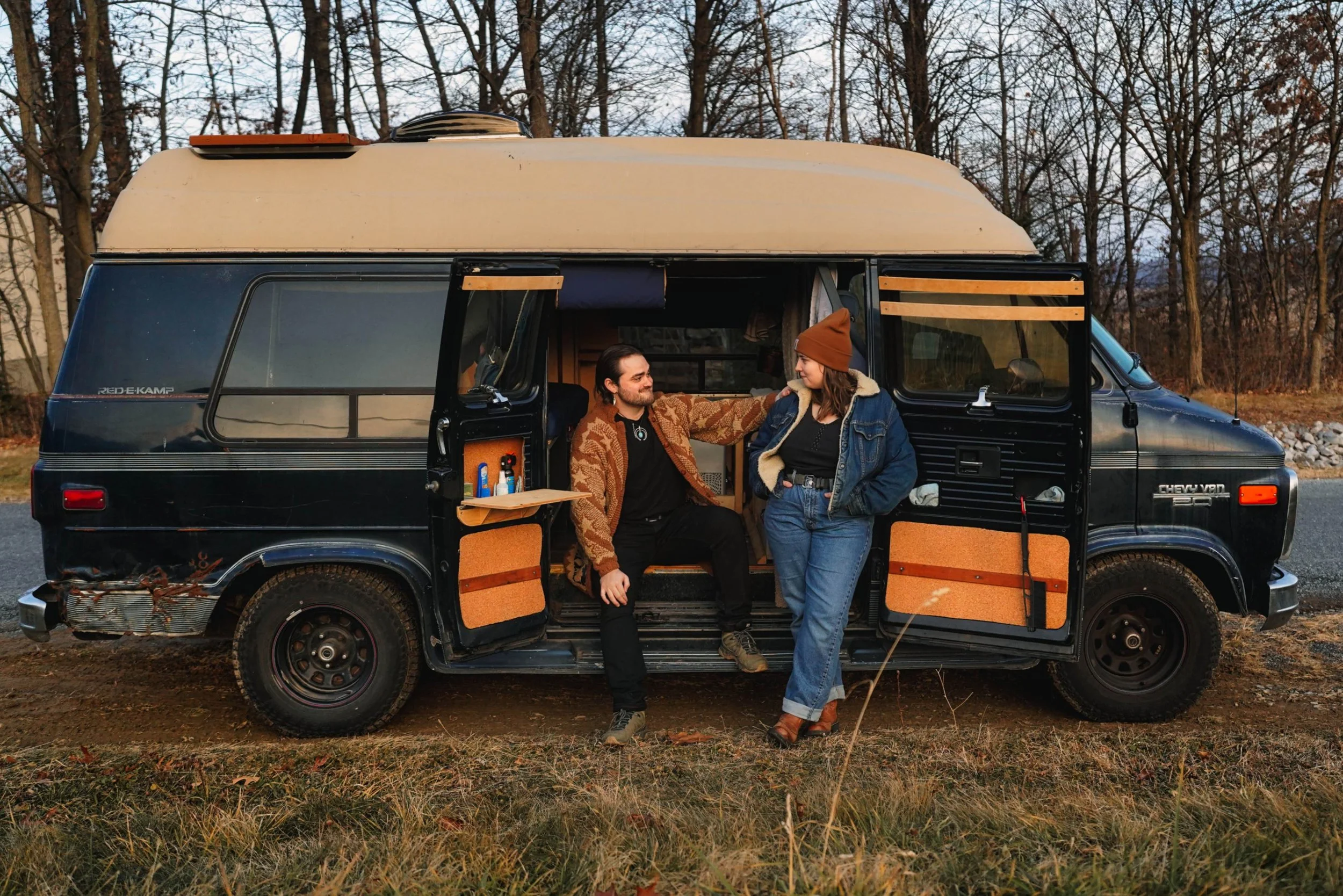 A man and woman standing beside a black camper van in an outdoor setting with trees in the background. The man is sitting on the van's open sliding door, and the woman is standing with her hands in her pockets, wearing a denim jacket and brown beanie