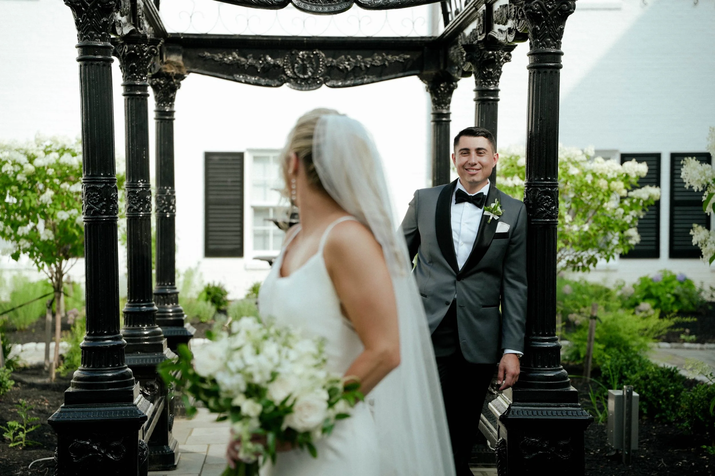 A wedding couple stands outdoors near a black decorative arch. The bride, in a white wedding dress and veil, holds a bouquet of white flowers. The groom, in a gray tuxedo with a black bow tie and boutonniere, smiles at her.