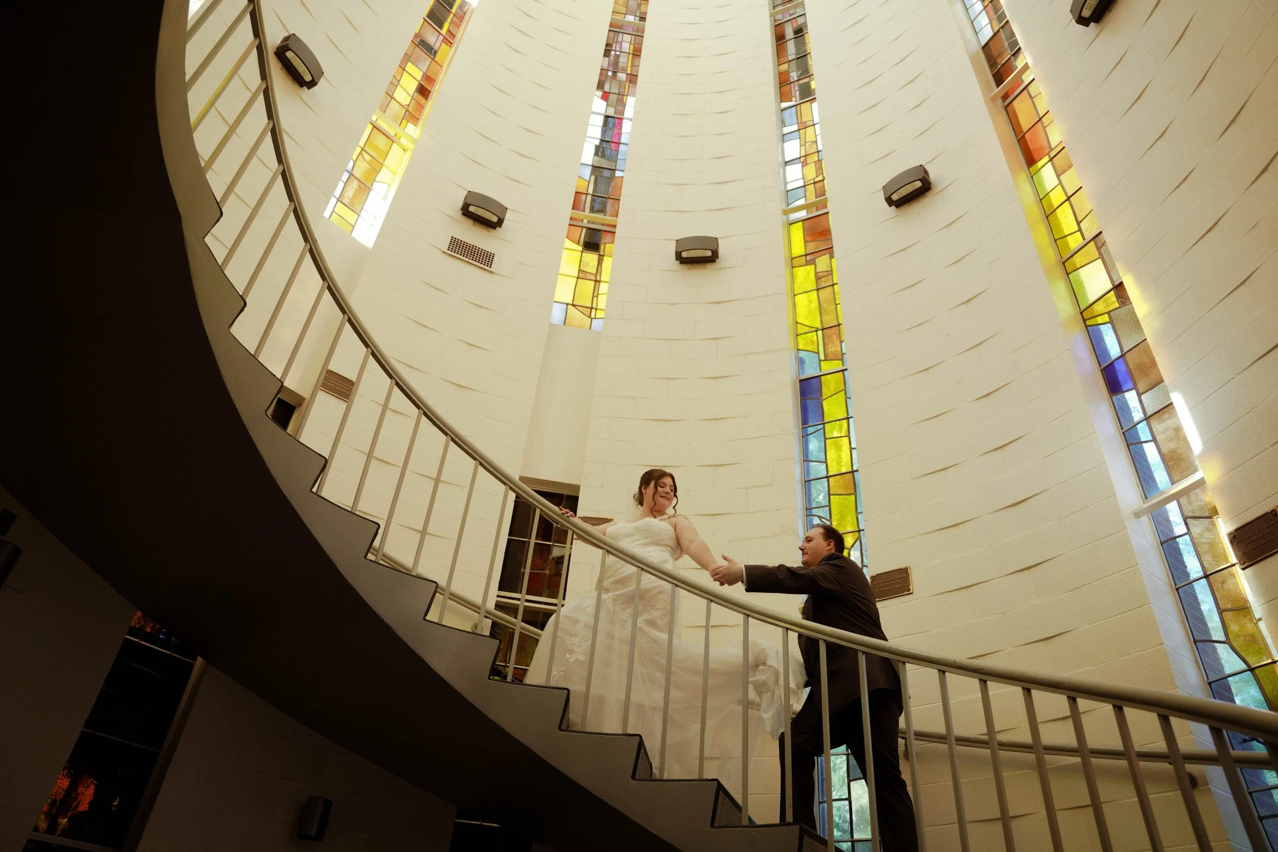 A bride and groom dancing on a curved staircase inside a church with tall stained glass windows.