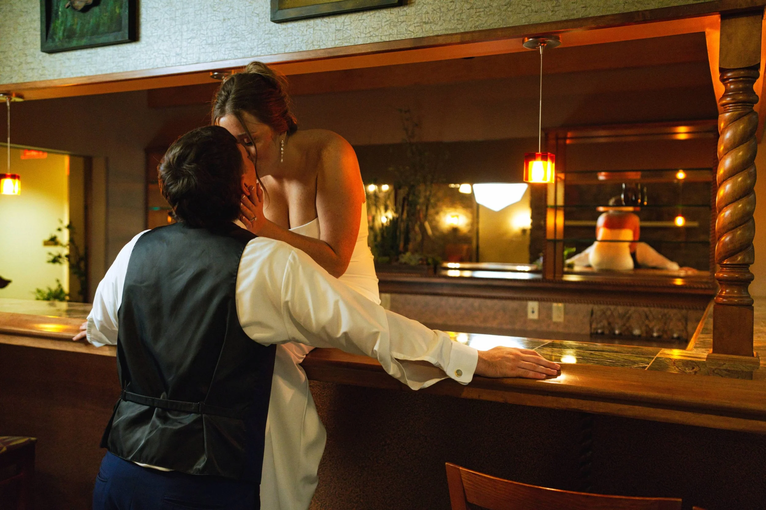 A woman in a white dress and a man in a vest sharing an intimate moment at a restaurant or bar, with the woman leaning over the counter towards the man, who is seated. The setting has warm lighting and a mirror in the background.