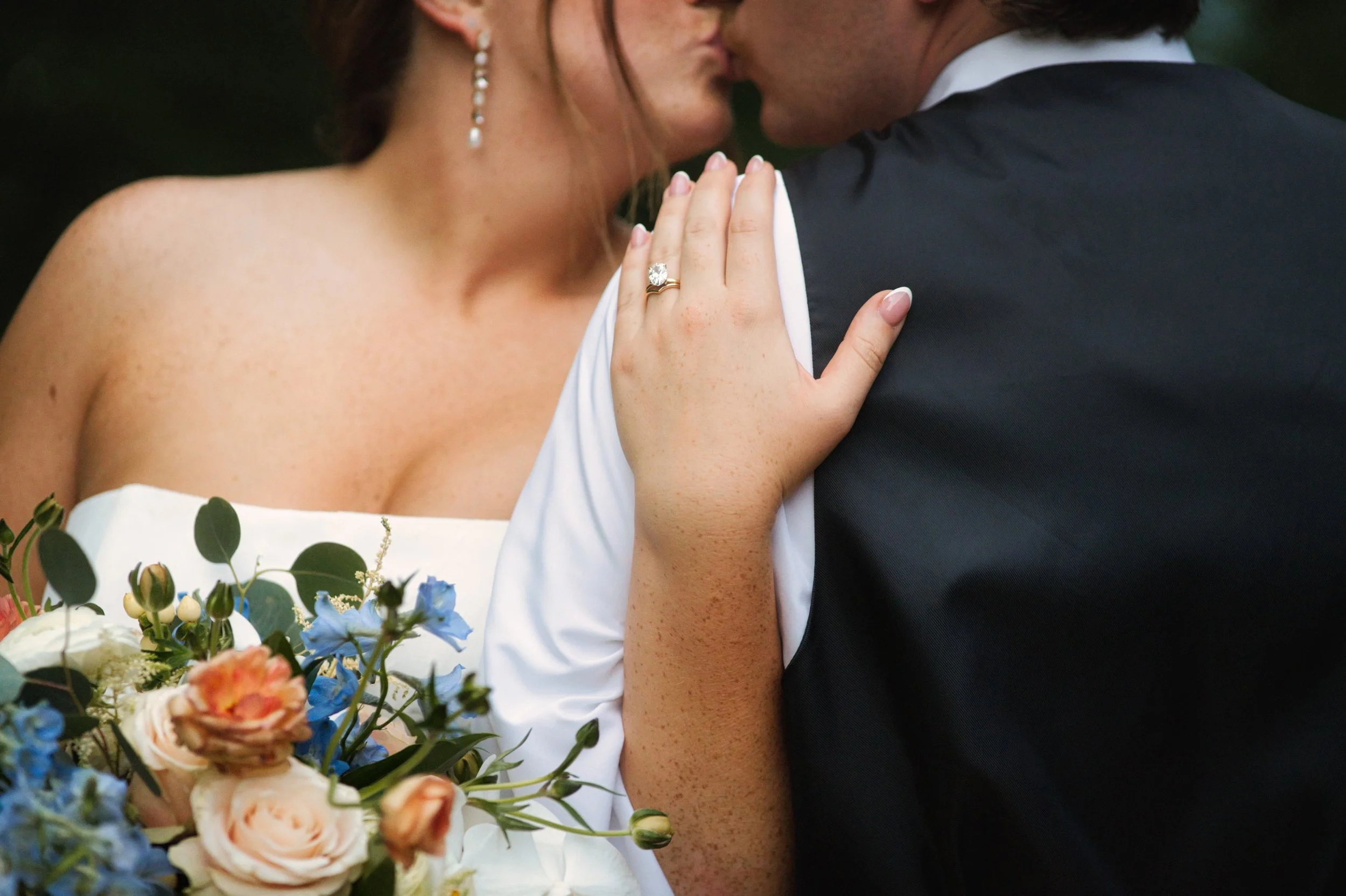 A bride and groom share a kiss, with the bride's hand, showing an engagement ring, resting on the groom's shoulder. The bride has freckles and wears earrings, and the groom is dressed in a dark suit. A bouquet of flowers, including roses and other bl