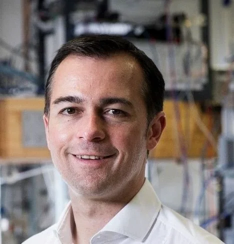 Portrait of a man with dark hair, wearing a white shirt, smiling in an indoor setting with blurred background.