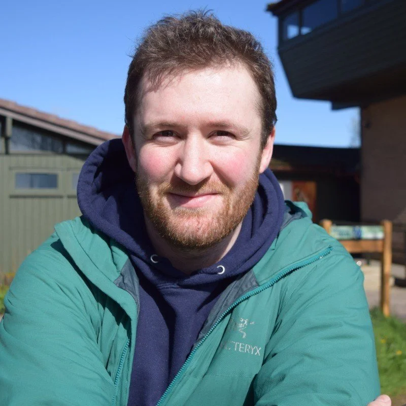 A man with a beard smiling outdoors on a sunny day, wearing a blue hoodie and a green jacket.