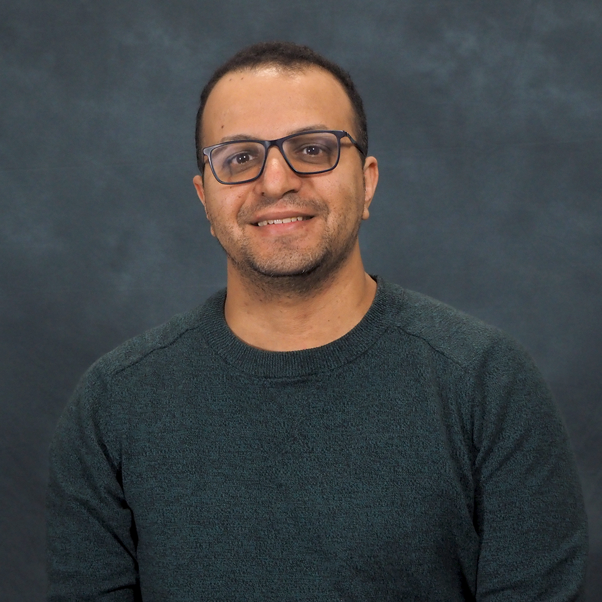 Headshot of a man with short dark hair, wearing glasses and a dark green sweater, smiling against a dark backdrop.
