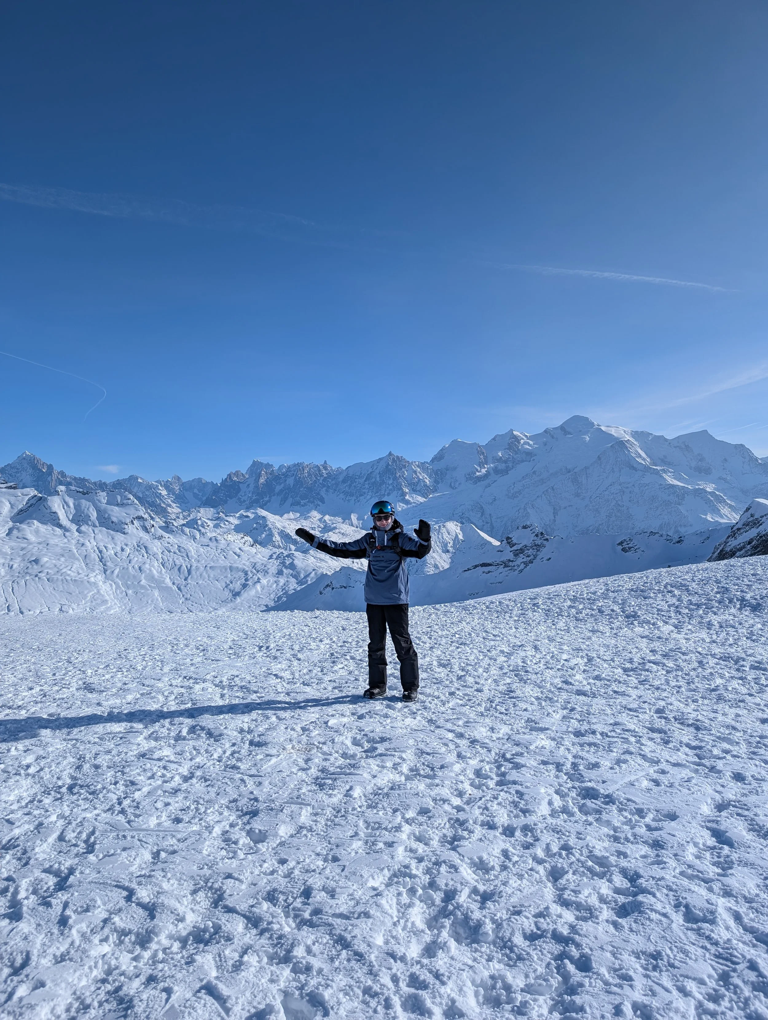 Person dressed in winter gear standing on snowy mountain landscape with snow-capped peaks and a clear blue sky in the background.