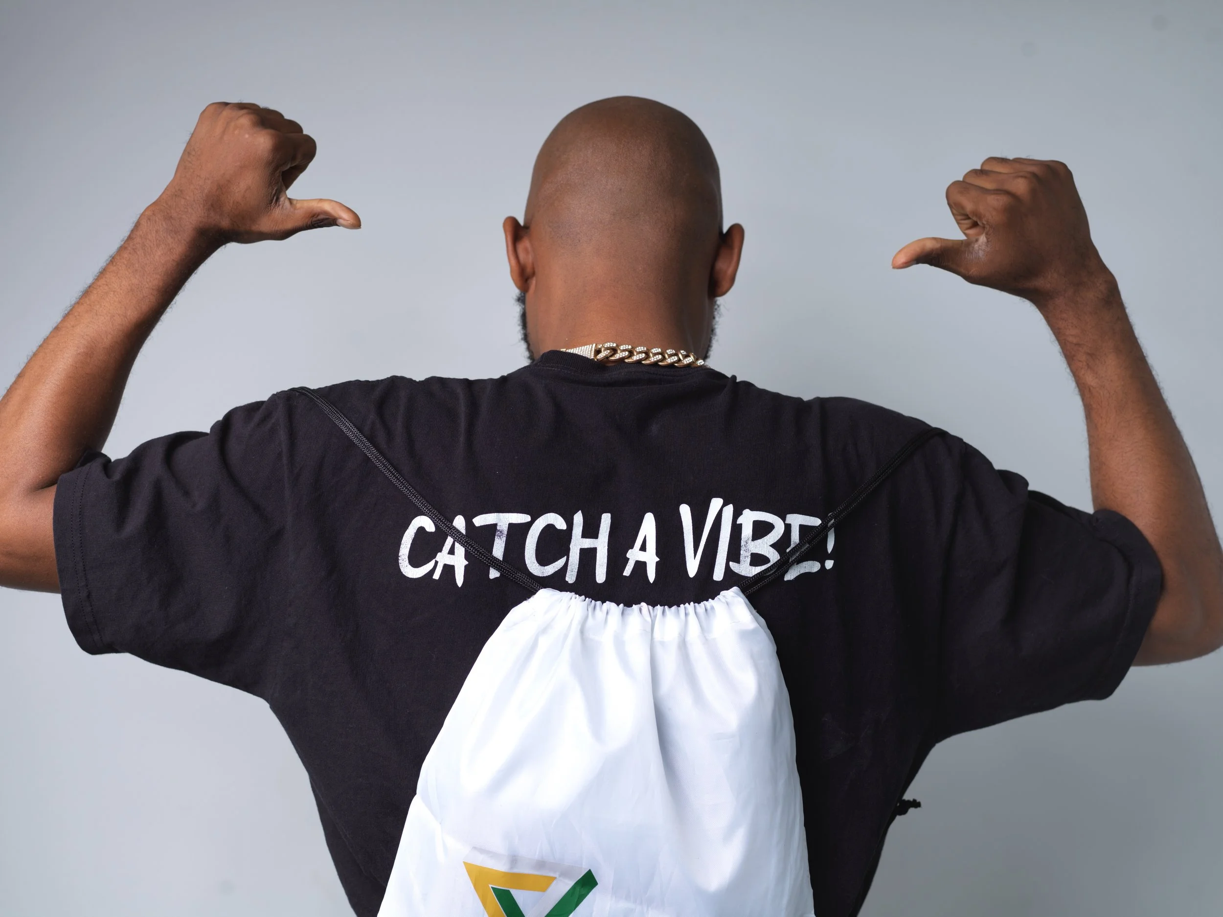 Back of a man wearing a black t-shirt with white text reading 'Catch a Vibe' and a white backpack. The man is flexing his muscles with both arms raised against a plain background.