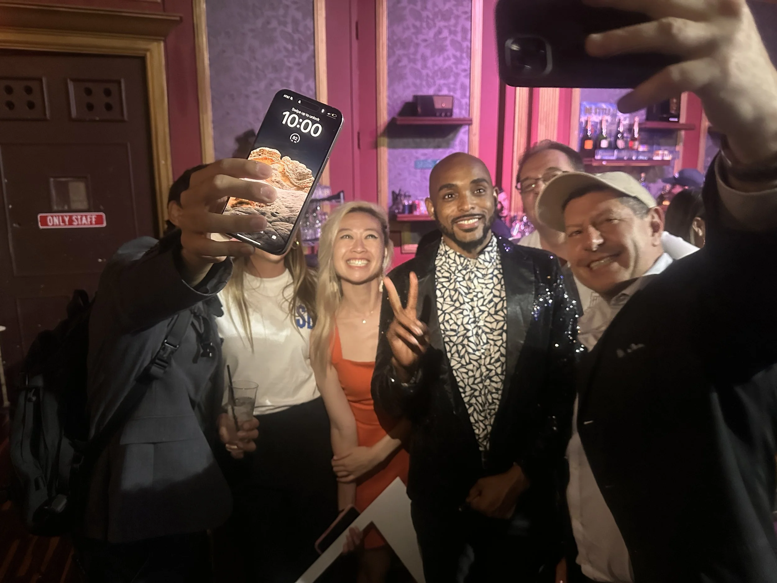 A group of people taking a selfie at a social gathering in a dimly lit room, with one person holding a smartphone in the foreground and smiling individuals in the background.