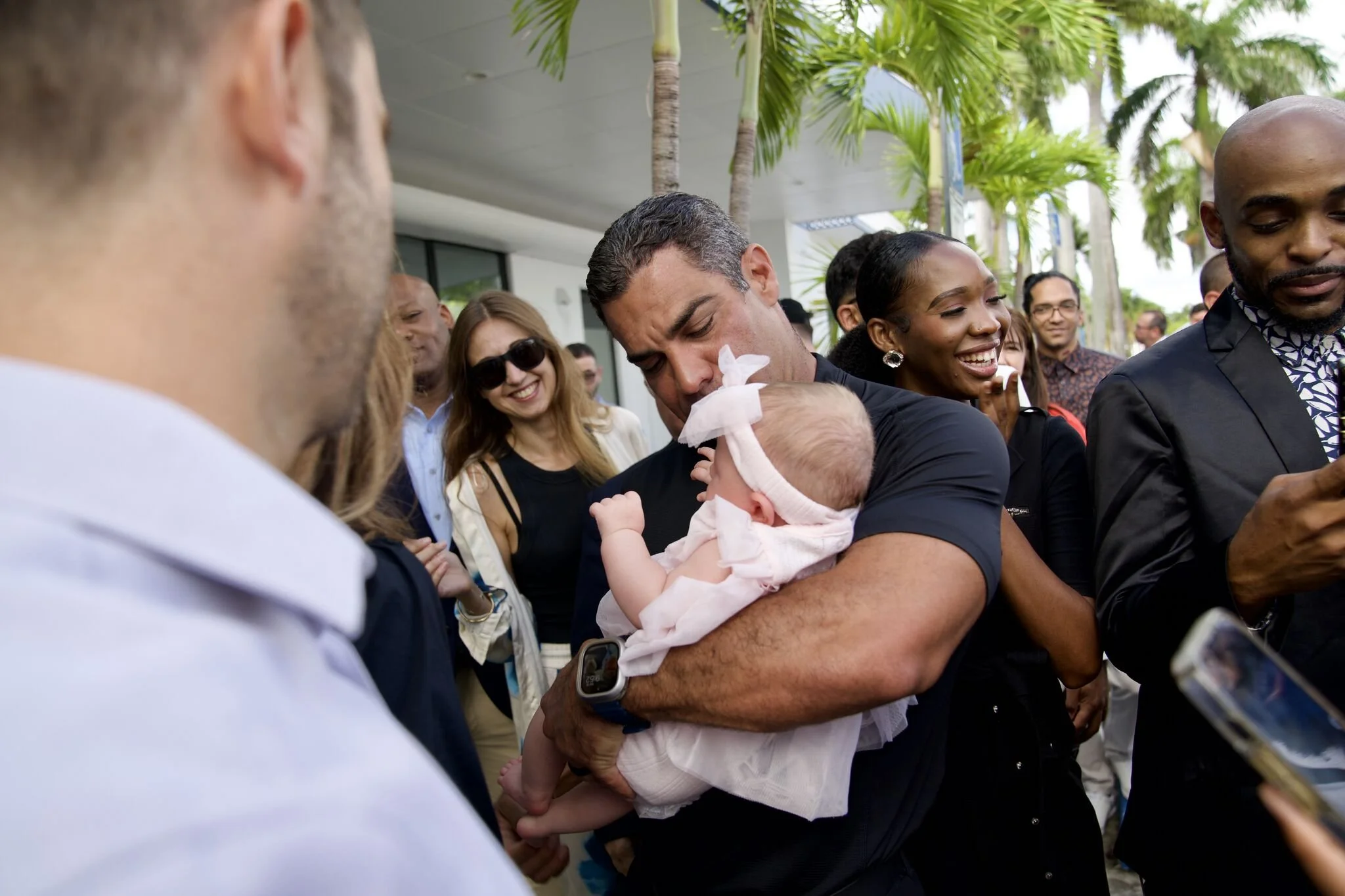A man holding a smiling baby girl wrapped in a white blanket, outdoors with palm trees and a crowd of smiling people around him.