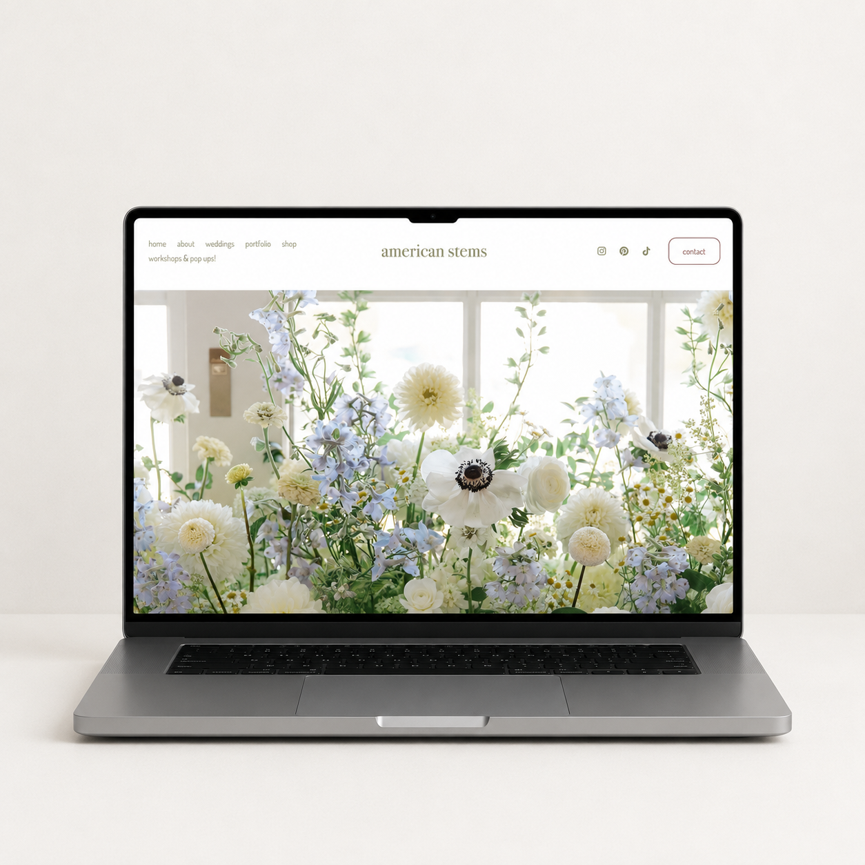 A laptop on a wooden desk displaying a website, with a glass vase of pink roses, a small terracotta pot with pencils, a notepad, and a large metal bucket with white and pink flowers in the background.