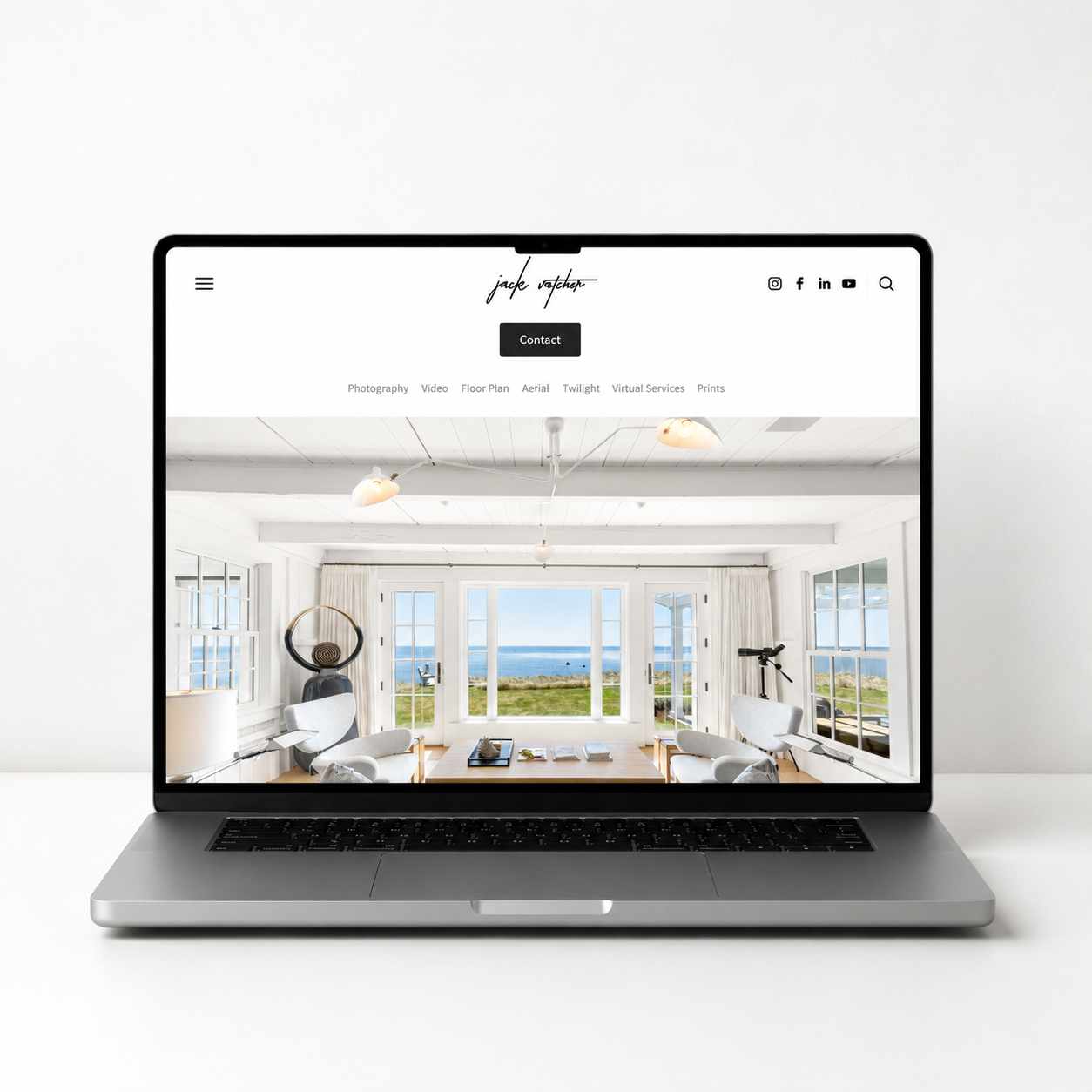 A laptop on a wooden desk displays a photo of a bright, airy room with large windows overlooking the ocean. To the left of the laptop is a black camera with a lens, and to the right is a black camera lens. In front of the laptop are three printed photographs of interior and exterior architecture.