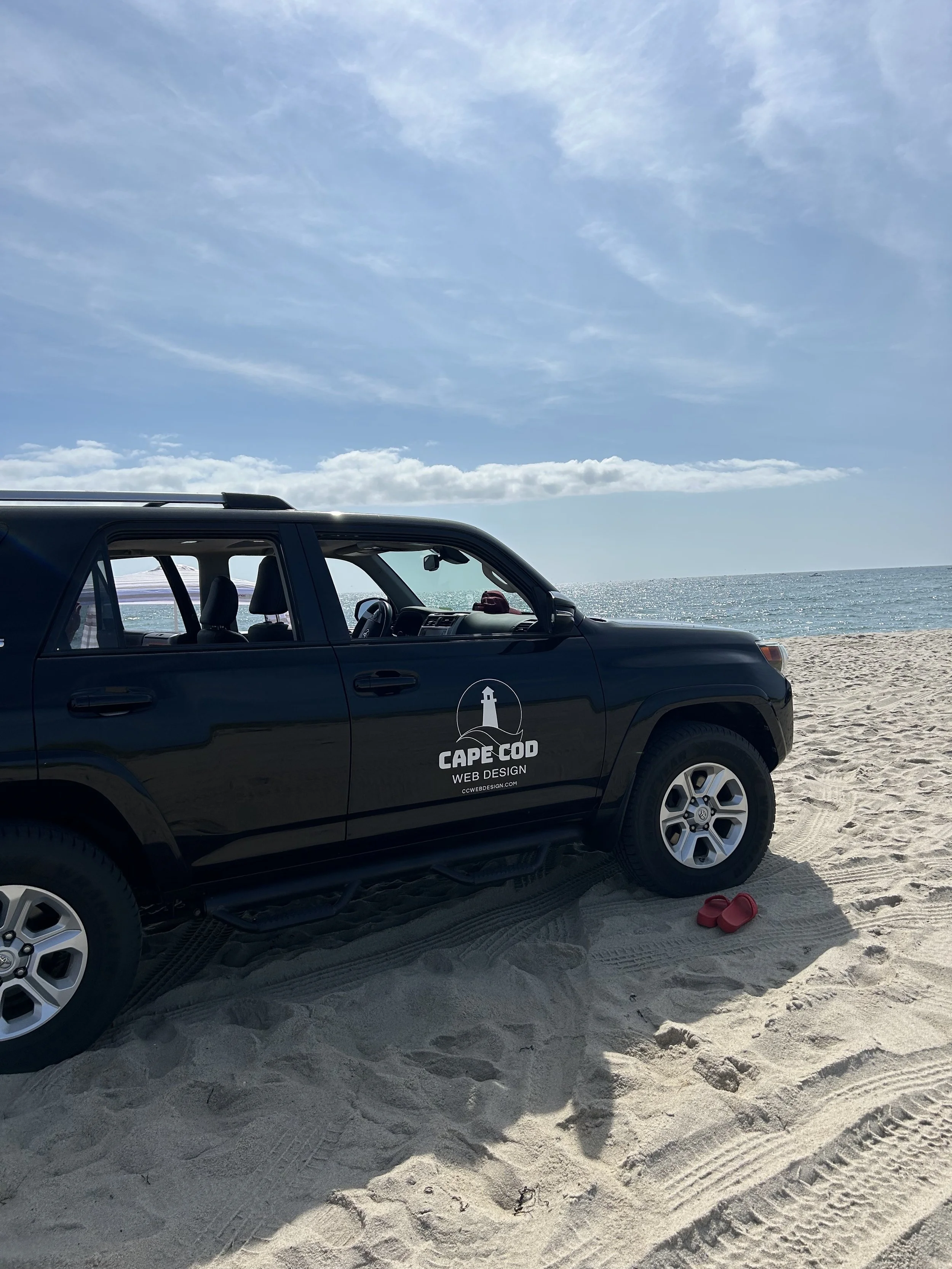 Black SUV parked on sandy beach with Cape Cod web design logo on the door, red sandals on the sand, and ocean with sky in the background.