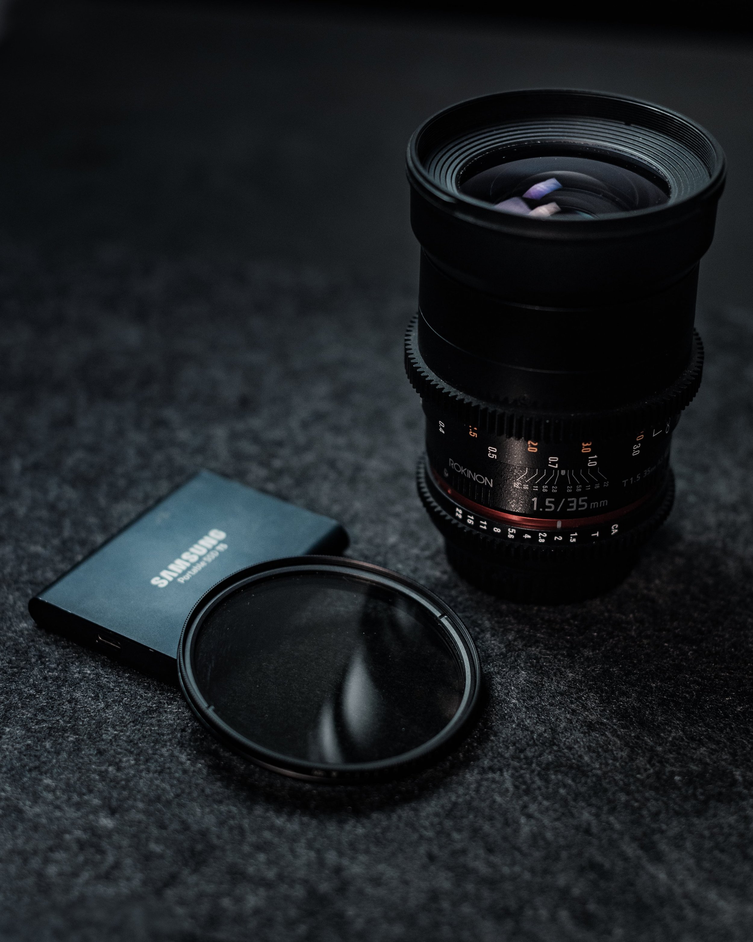Product Photo of a camera lens, circular filter, Samsung portable SSD on dark surface.