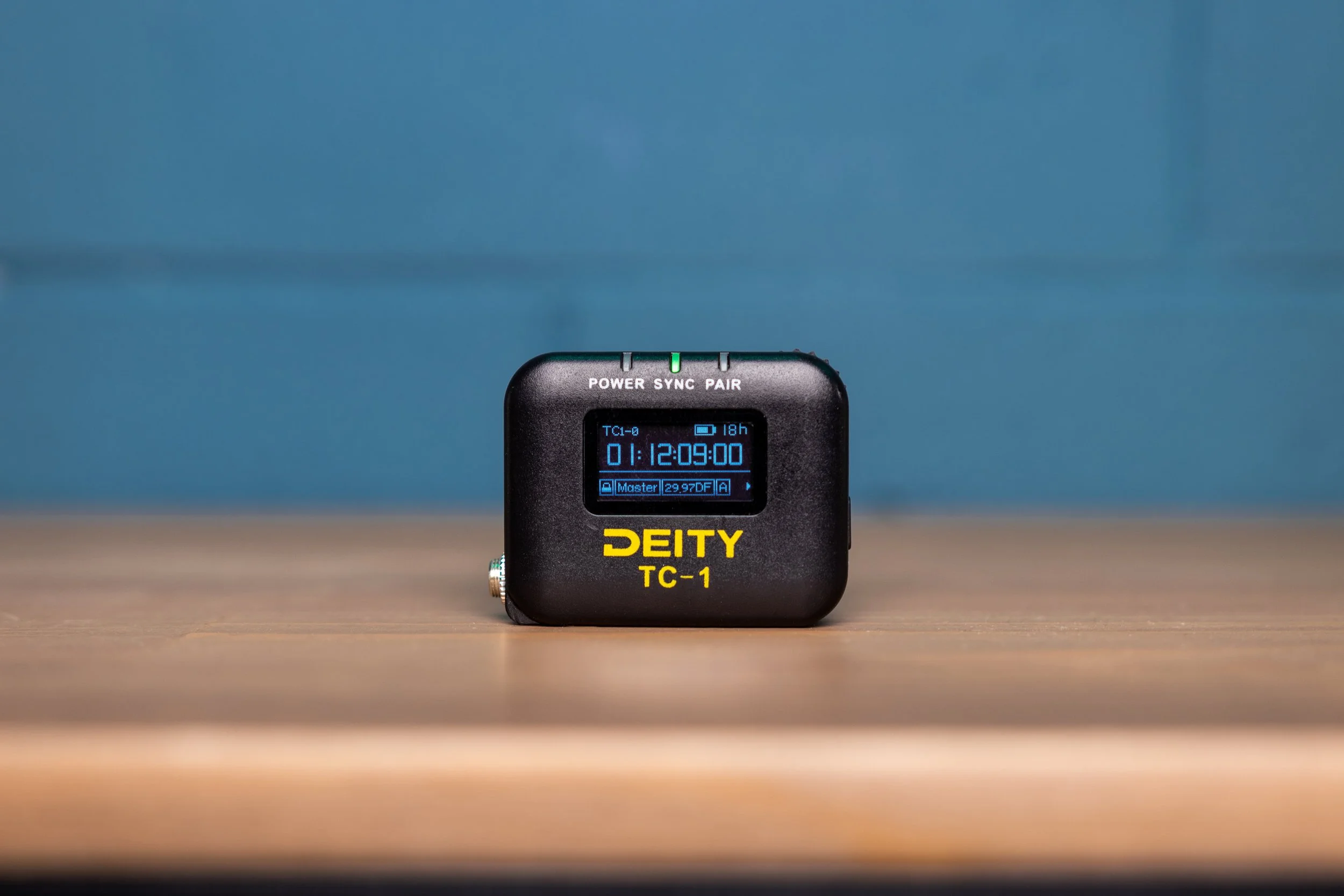 Product Photo of Deity TC-1 timecode generator on a wooden surface with a blue background, displaying sync information on its screen.
