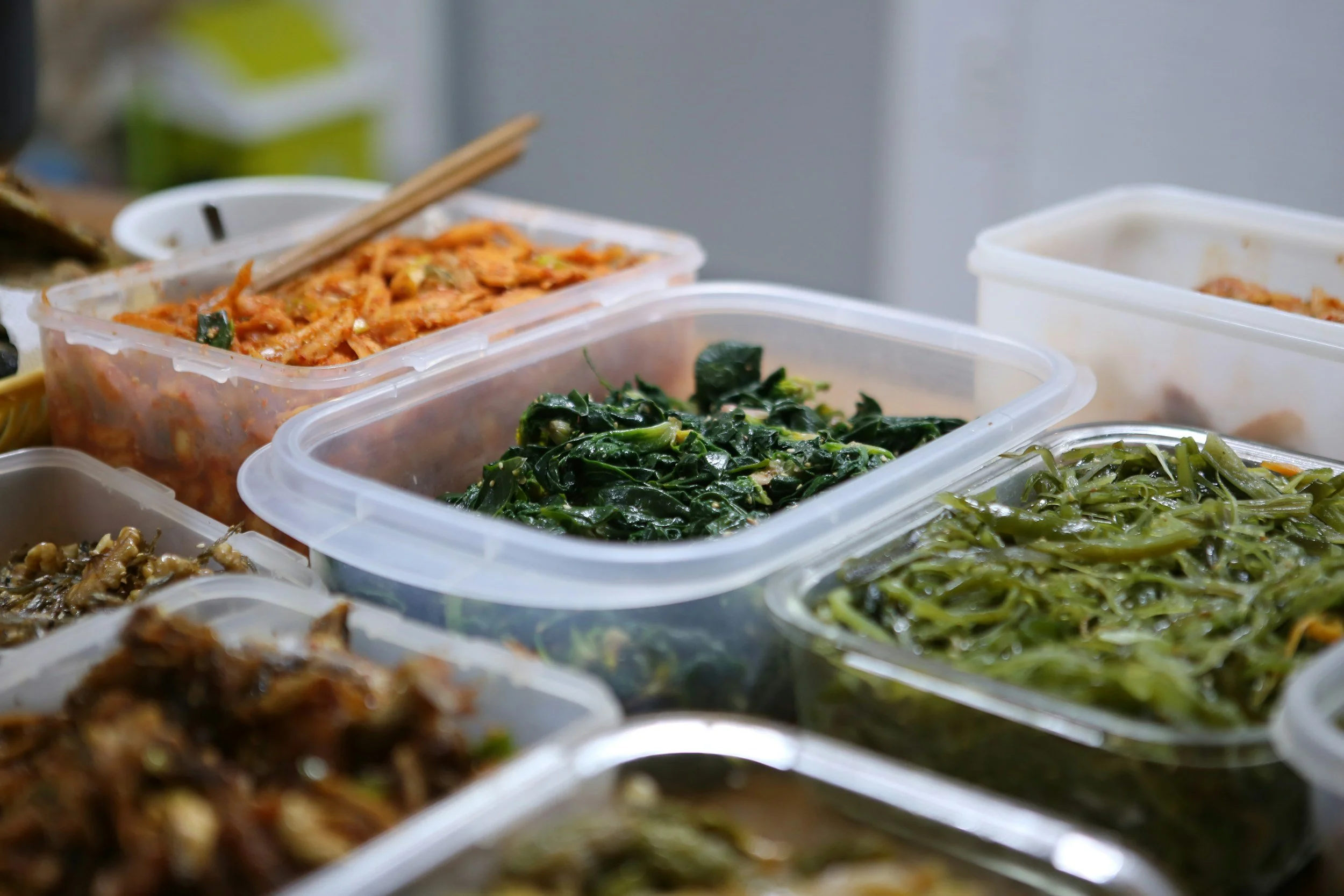 Close-up of various Asian side dishes in plastic containers, including kimchi, cooked greens, and seaweed, with chopsticks resting on one container.