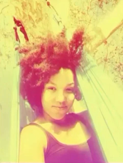 A person smiling while relaxing in a hammock, surrounded by trees and bright sunlight, with a tinted color effect.