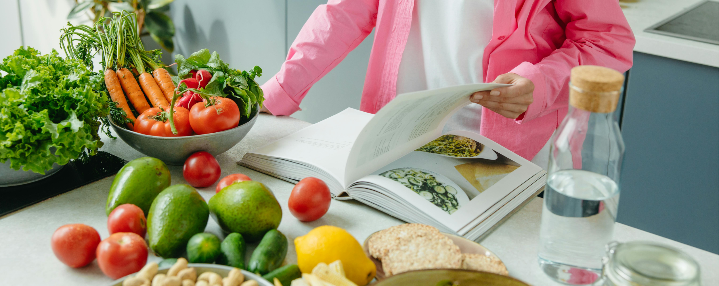Woman standing at kitchen counter with fresh vegetables and reading a cookbook