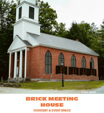 Brick church with white steeple and black-shuttered windows, surrounded by green trees.