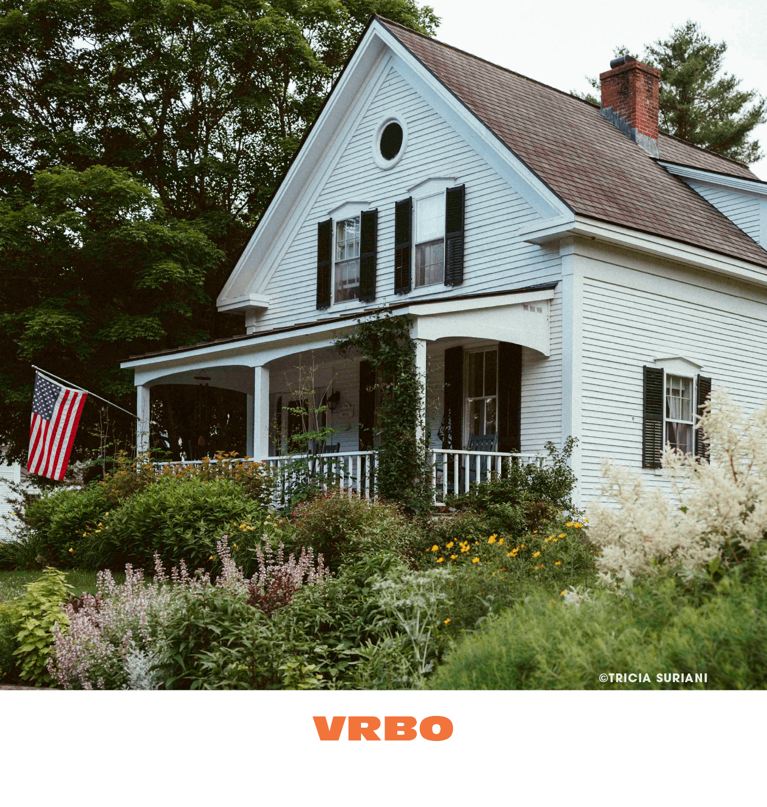 A white two-story house with black shutters, a porch with white railing, an American flag, and a garden with various flowers.