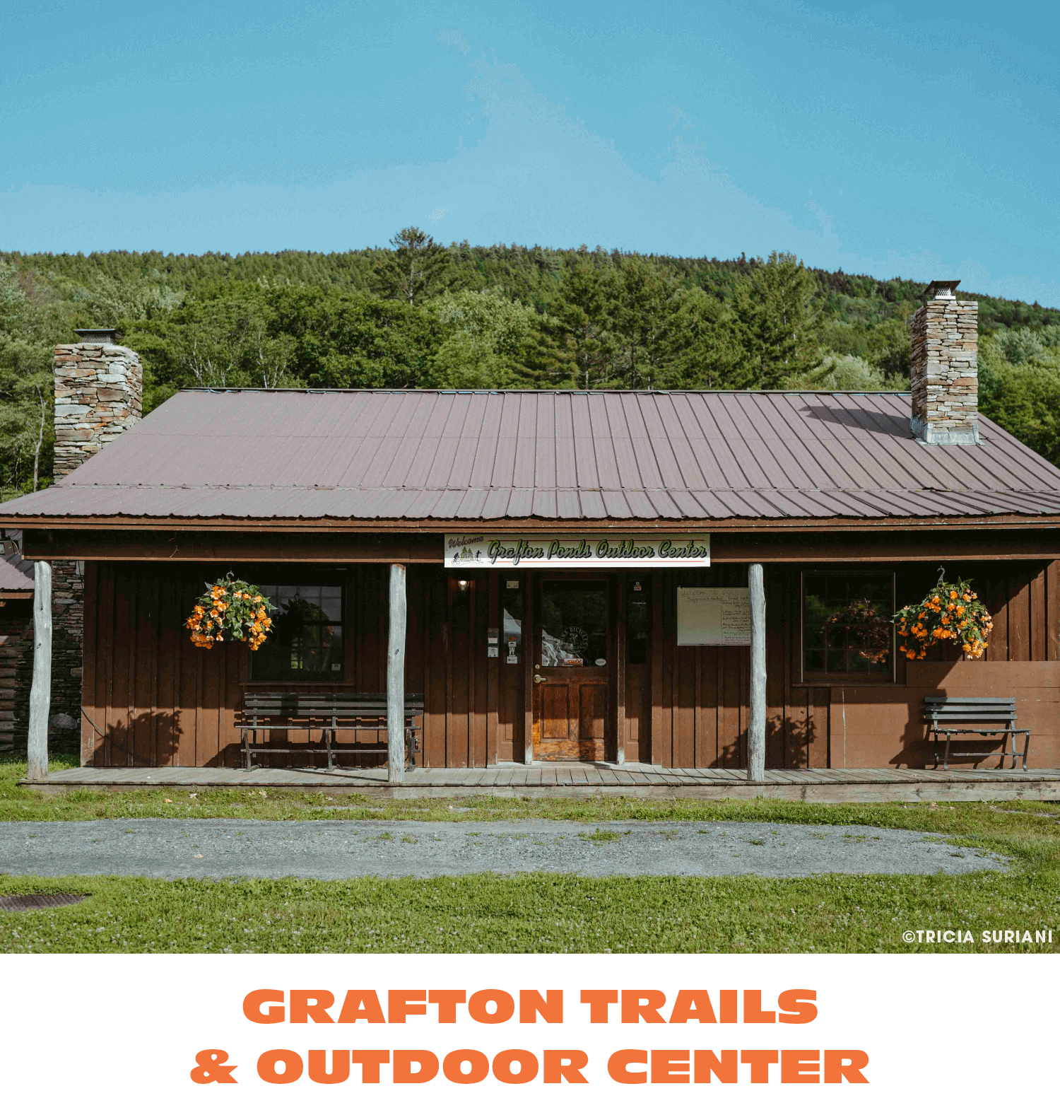 A rustic wooden building with a metal roof and two stone chimneys, surrounded by green trees and grass, with hanging flower baskets and a sign that reads 'Grafton Trails & Outdoor Center'.