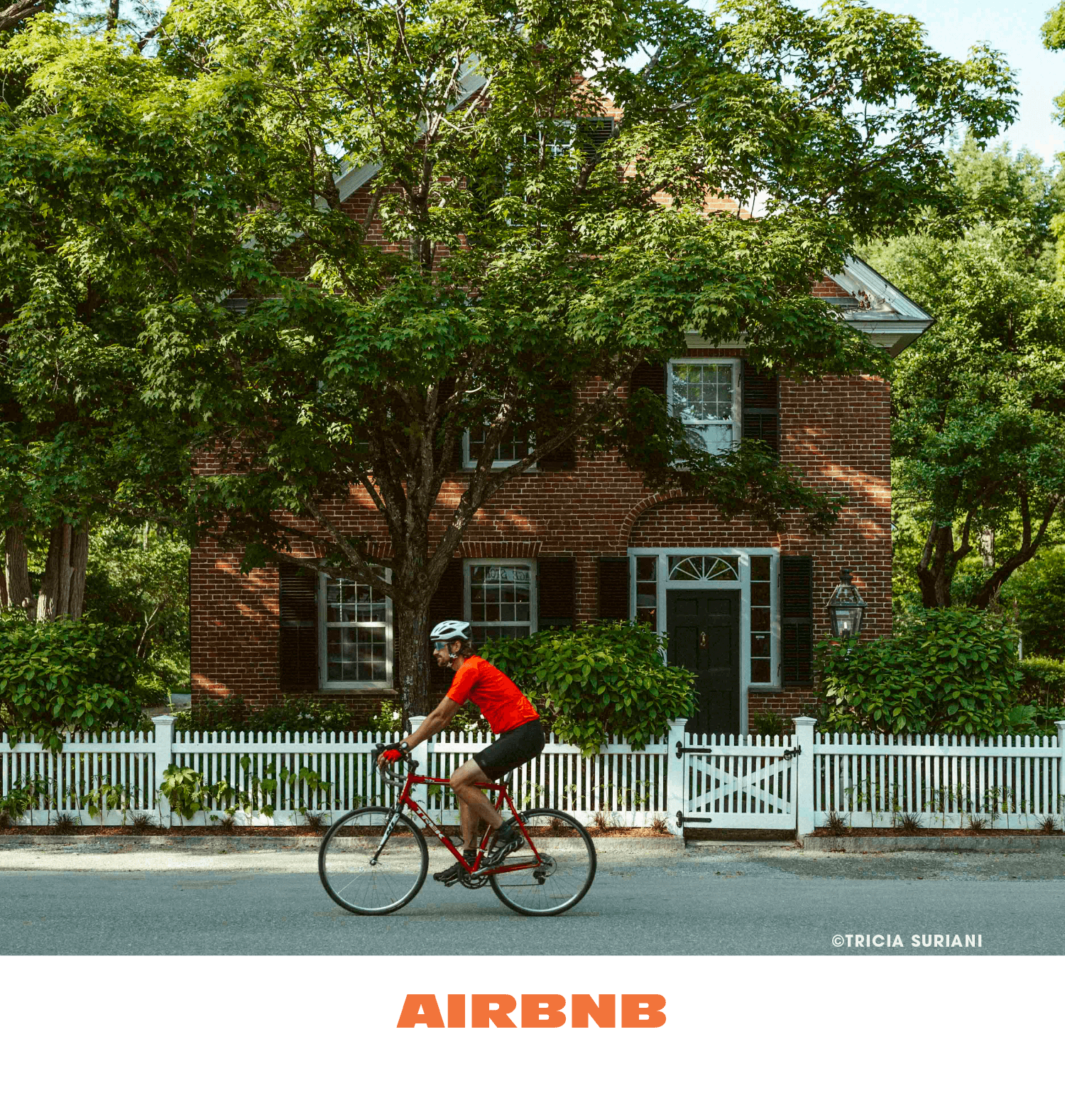 A person in a red shirt and black shorts riding a bicycle past a brick house with a white picket fence and green trees.