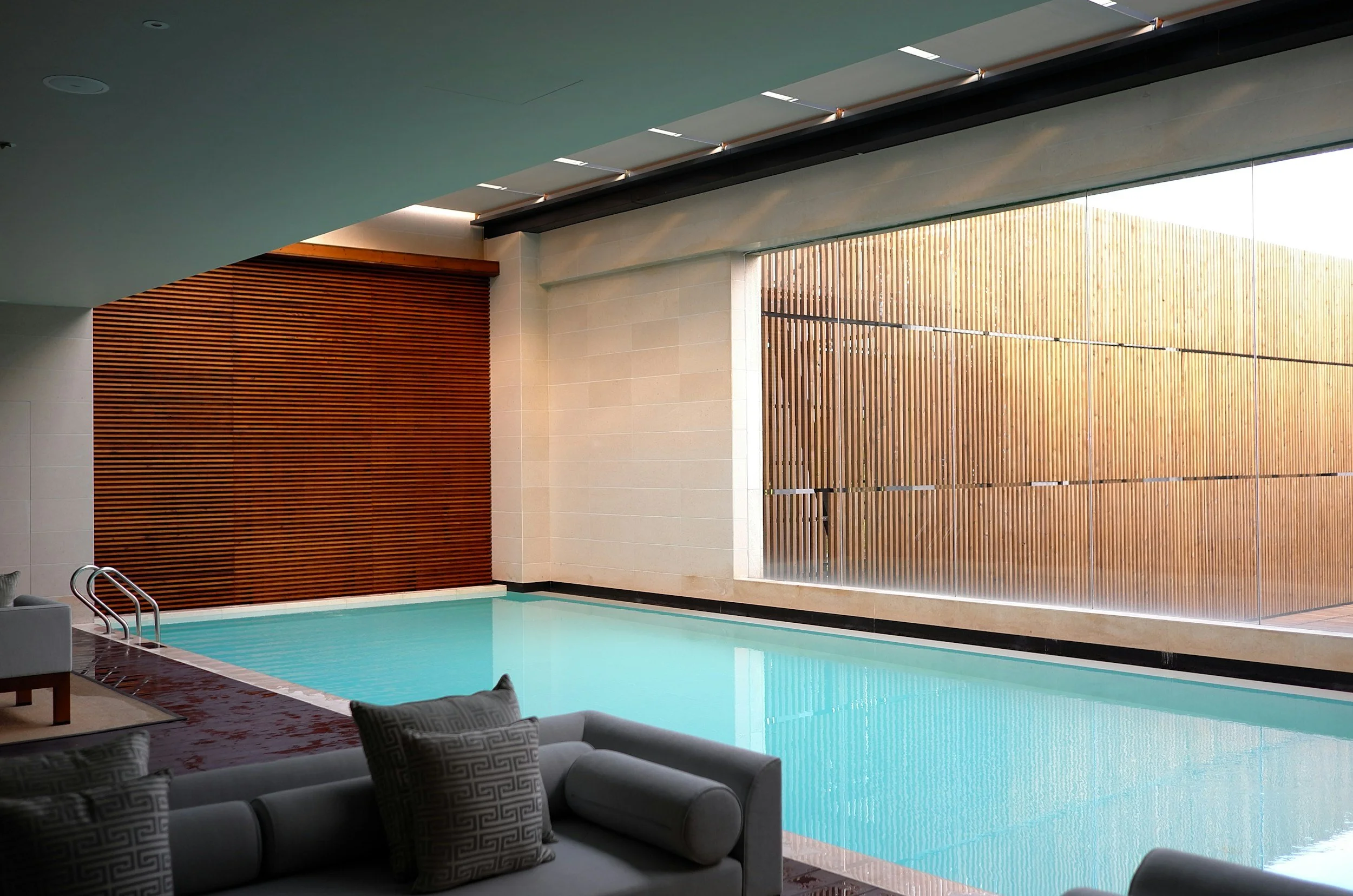 Indoor swimming pool with modern wooden wall panels and large windows, furnished with a gray sofa and pillows.