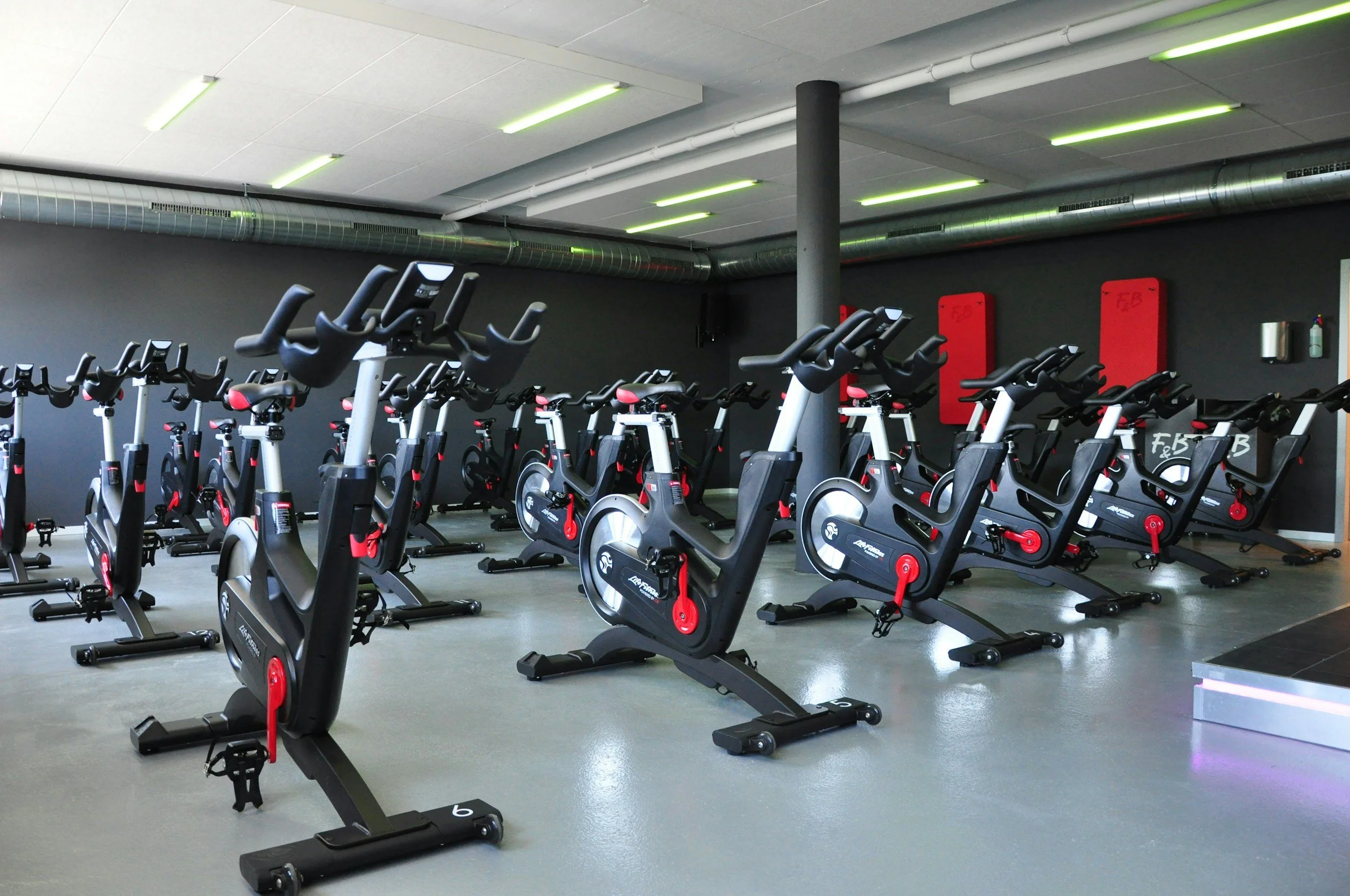 Indoor gym with multiple stationary exercise bikes arranged in rows, black walls, and ceiling lights.
