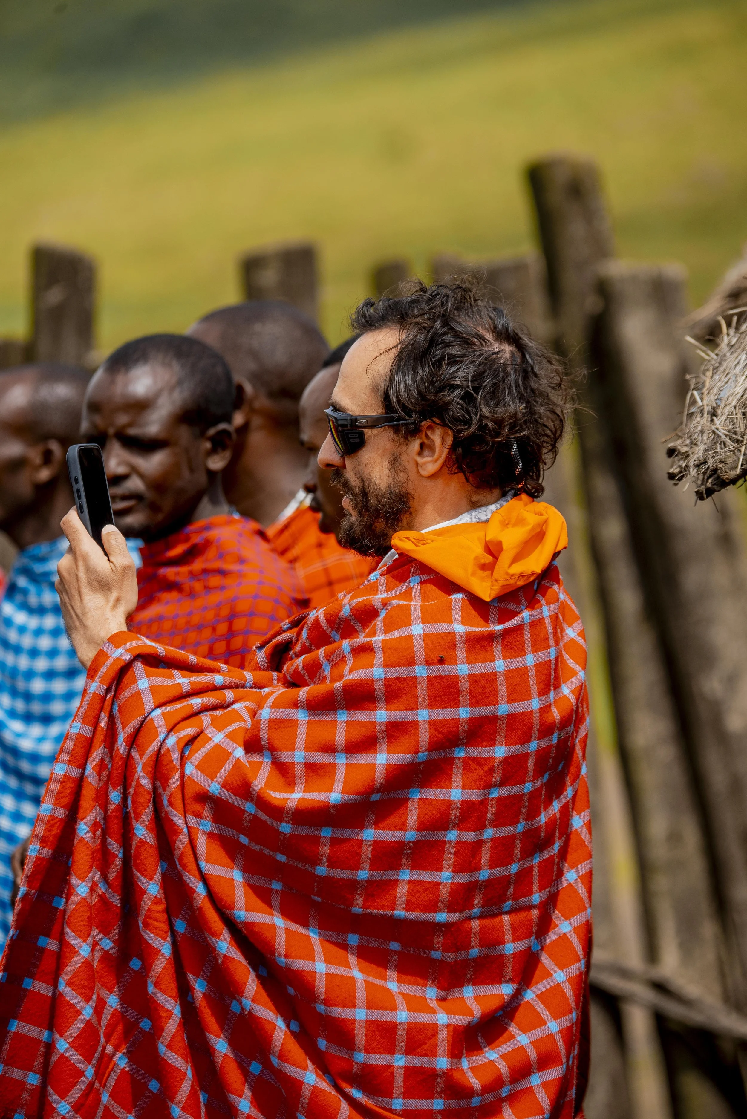 Man with sunglasses and curly hair in traditional Maasai blanket taking a photo with a smartphone among Maasai people in similar attire.