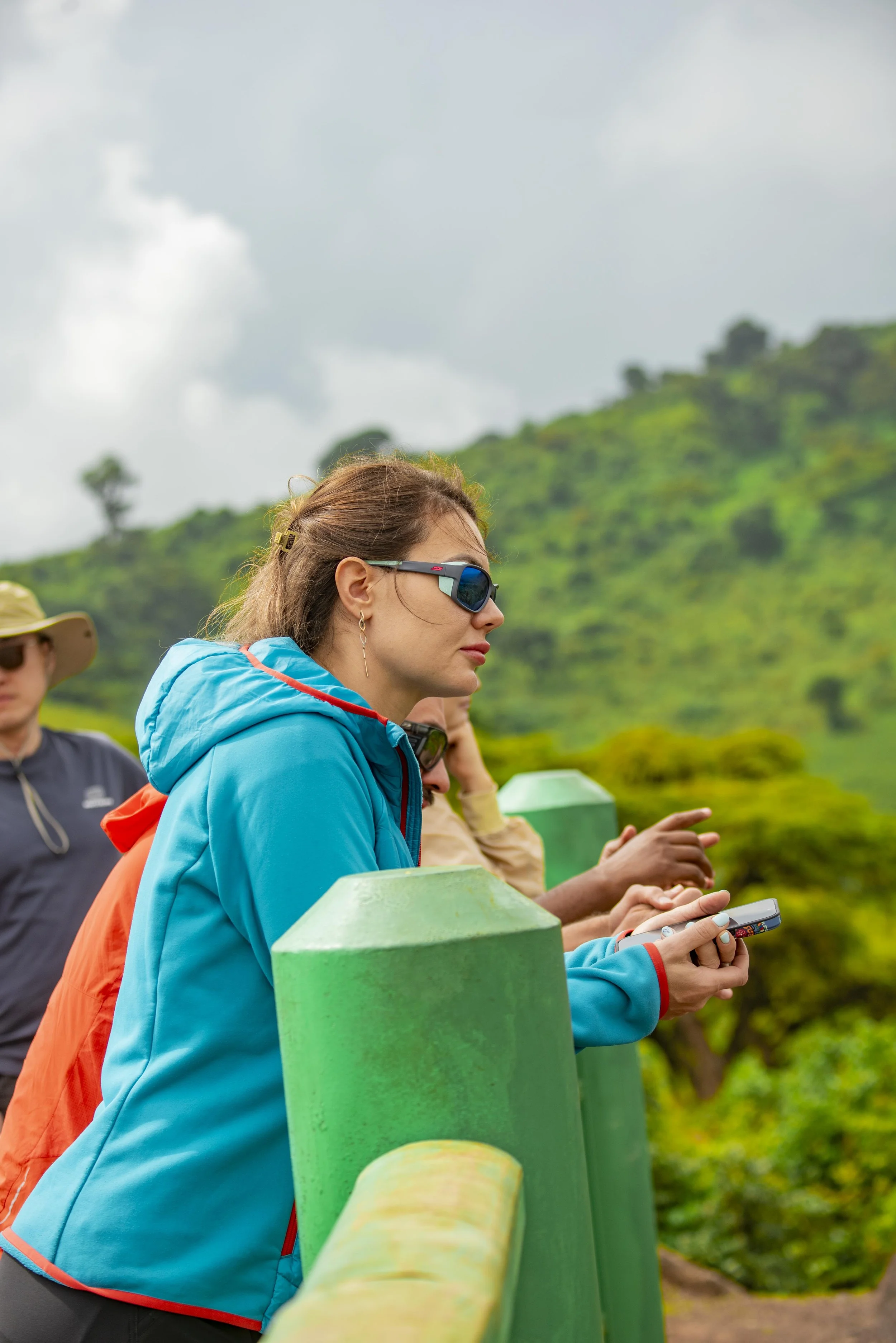 Three women standing outdoors on a cloudy day near green mountain scenery, wearing outdoor jackets and sunglasses, with one woman holding a smartphone.