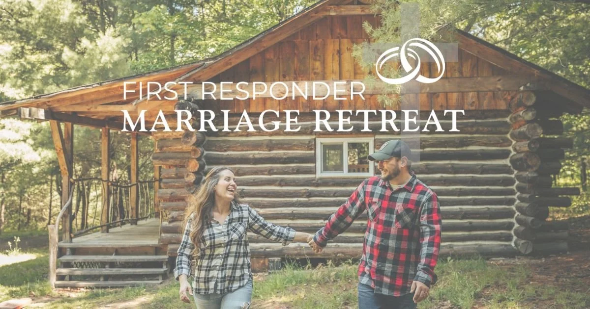A couple holding hands and smiling in front of a log cabin surrounded by trees, advertising a marriage retreat for first responders.